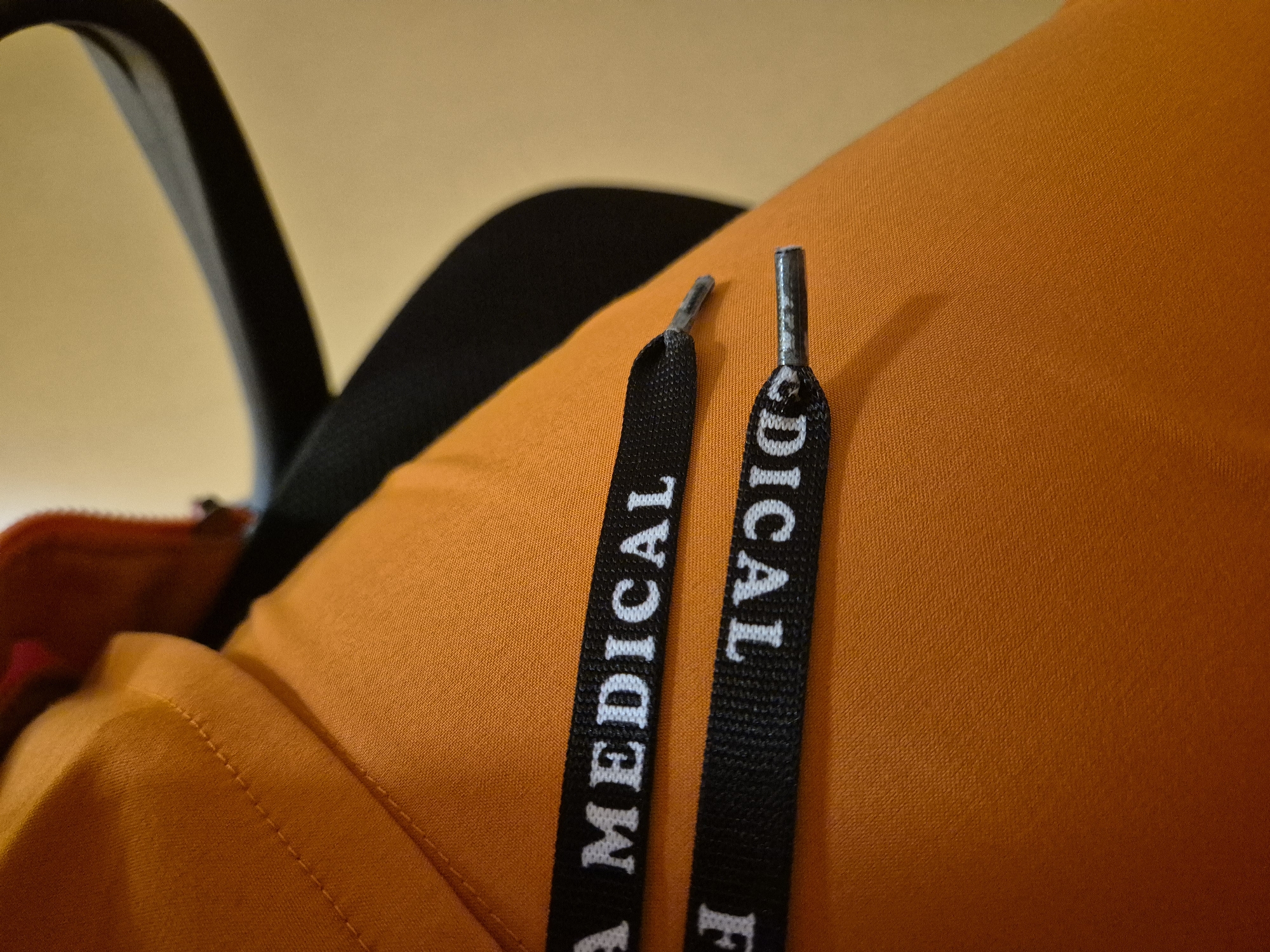 The image shows a close-up view of a bright orange garment, possibly a jacket or hoodie, with two black drawstrings featuring white lettering. The top drawstring reads "A MEDICAL," with the "A" partially visible. The bottom drawstring is reversed and reads "ICAL F," with "ICAL" clearly legible. The drawstrings have silver tips. A portion of a black chair is visible in the background, out of focus. The overall setting is a neutral, indoor environment.