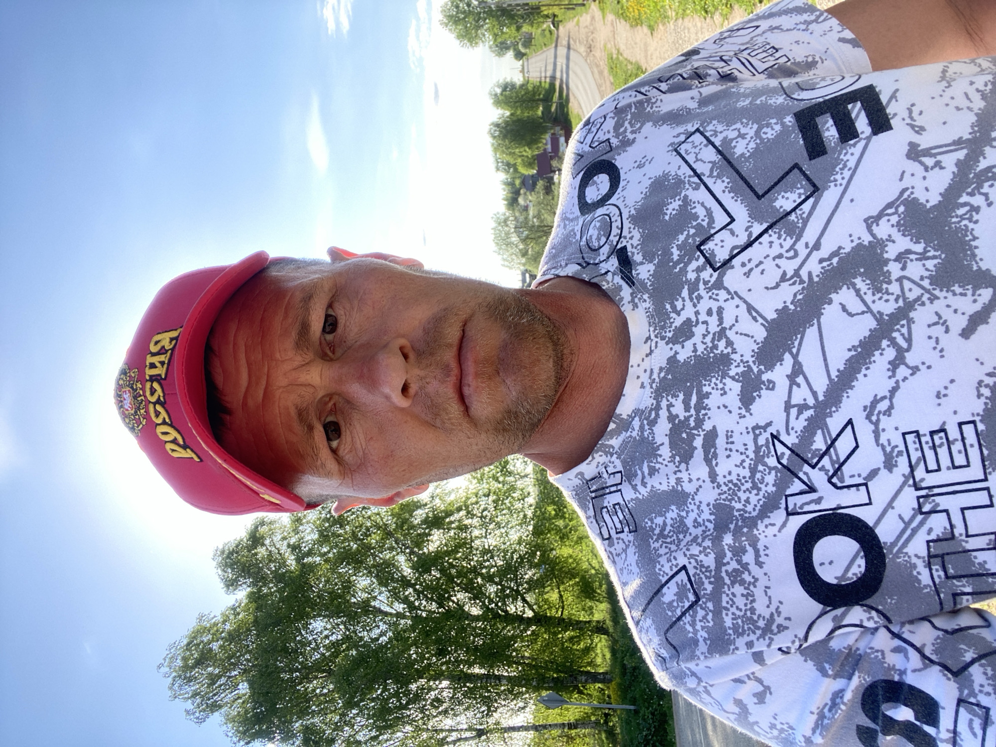 A middle-aged man stands outdoors in the daytime, wearing a white t-shirt with a grey and black patterned design and the words "LOOK LIKE" printed on it. He is also wearing a red baseball cap with gold lettering and a crest on the front. The man is facing the camera with a neutral expression. Behind him, there is green foliage and a street. The location is Фоки, Russia. The sun is shining brightly.