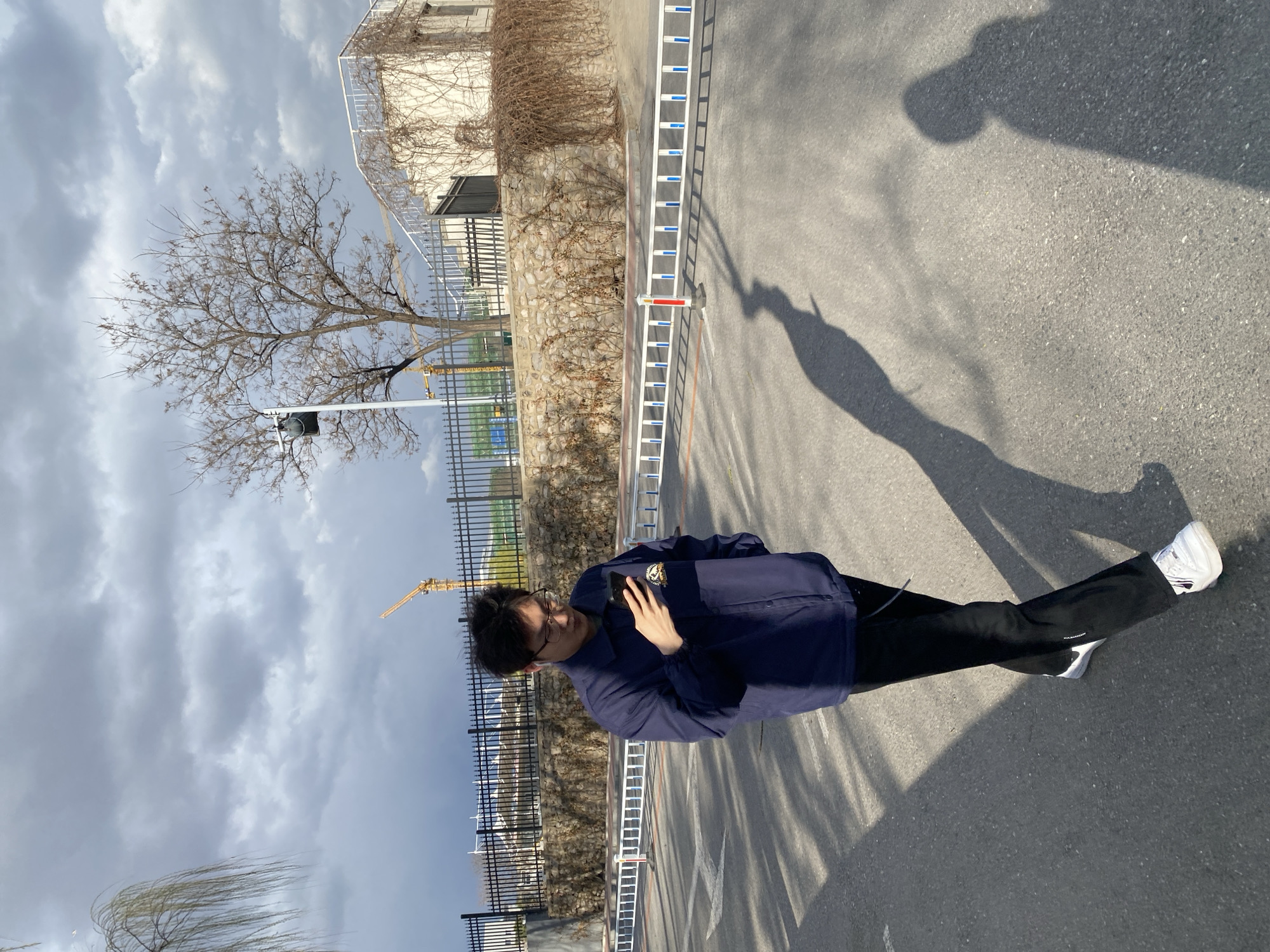 A young man stands on an asphalt road in what appears to be a public space, possibly a park or a street in the urban area of Zhongtun, China. The location exhibits a mix of natural and urban elements, including a building with ivy climbing on its wall, a bare tree, a crane in the distance, and a fence running along the roadside. The sky overhead is overcast. The man is wearing a navy blue jacket, black pants, and white sneakers, holding his phone. A long shadow is cast on the road surface.