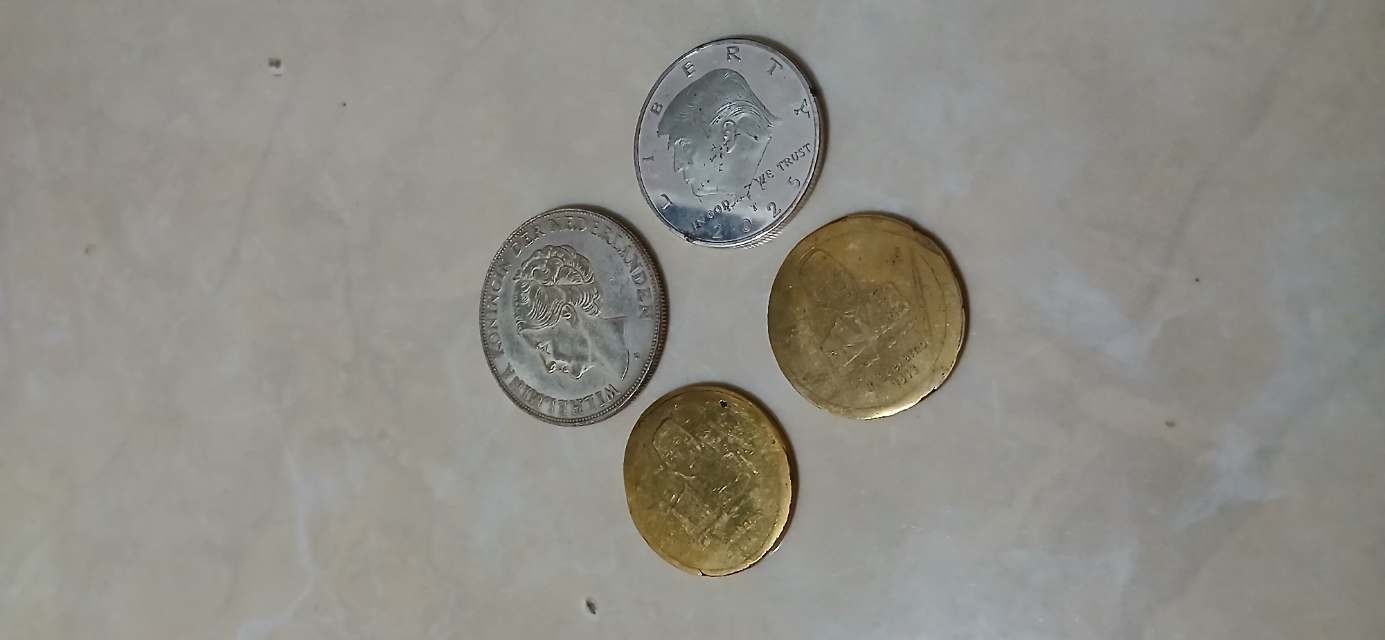 Four coins are arranged in a diamond shape on a light-colored, marble-patterned surface. The top coin is silver and features a profile of a woman with text around the edge. To the left and bottom are two gold-colored coins, each with an embossed image and text. To the right is another silver coin with a profile of a man and the words "WE TRUST" and "2025". The surface appears to be a countertop or floor with a subtle marble pattern. There are no people or animals visible in the image, and no specific location within Samarinda, Indonesia, can be identified from the visual information provided.
