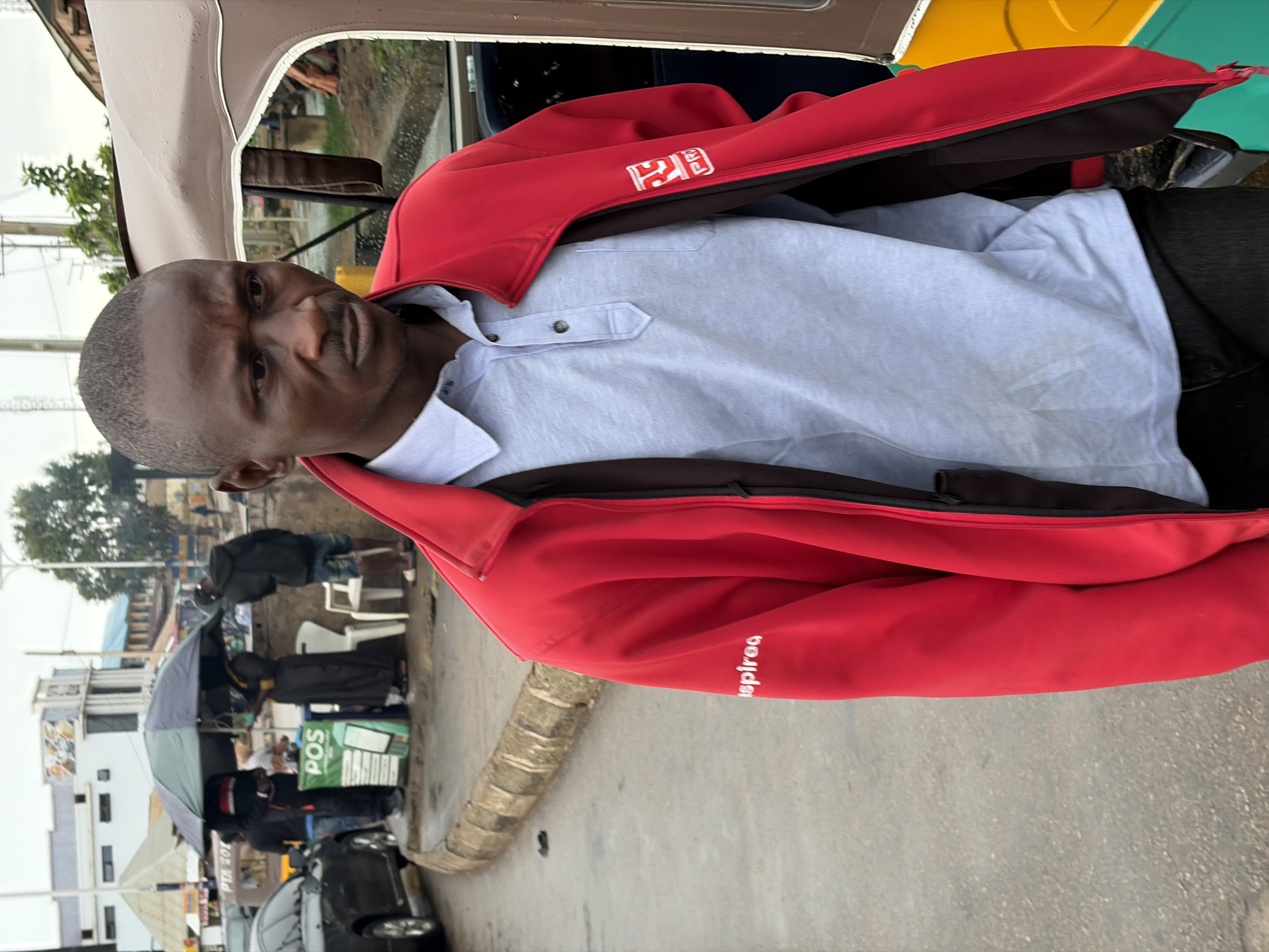 The image shows a man in the foreground wearing a red jacket over a light blue collared shirt. He has a serious expression and is looking towards the camera. The background is out of focus but appears to be an outdoor scene with some buildings and people. To the left of the man, there is a street with a few individuals, some under an umbrella, and a POS machine on a stand. A black car is parked on the street. The lighting suggests it is daytime, and the presence of an umbrella might indicate overcast or rainy weather. The man's red jacket has a logo on the left chest area and text on the sleeve that reads "inspiraa". The scene appears to be a street in Jos, Nigeria, possibly a commercial area with vendors.