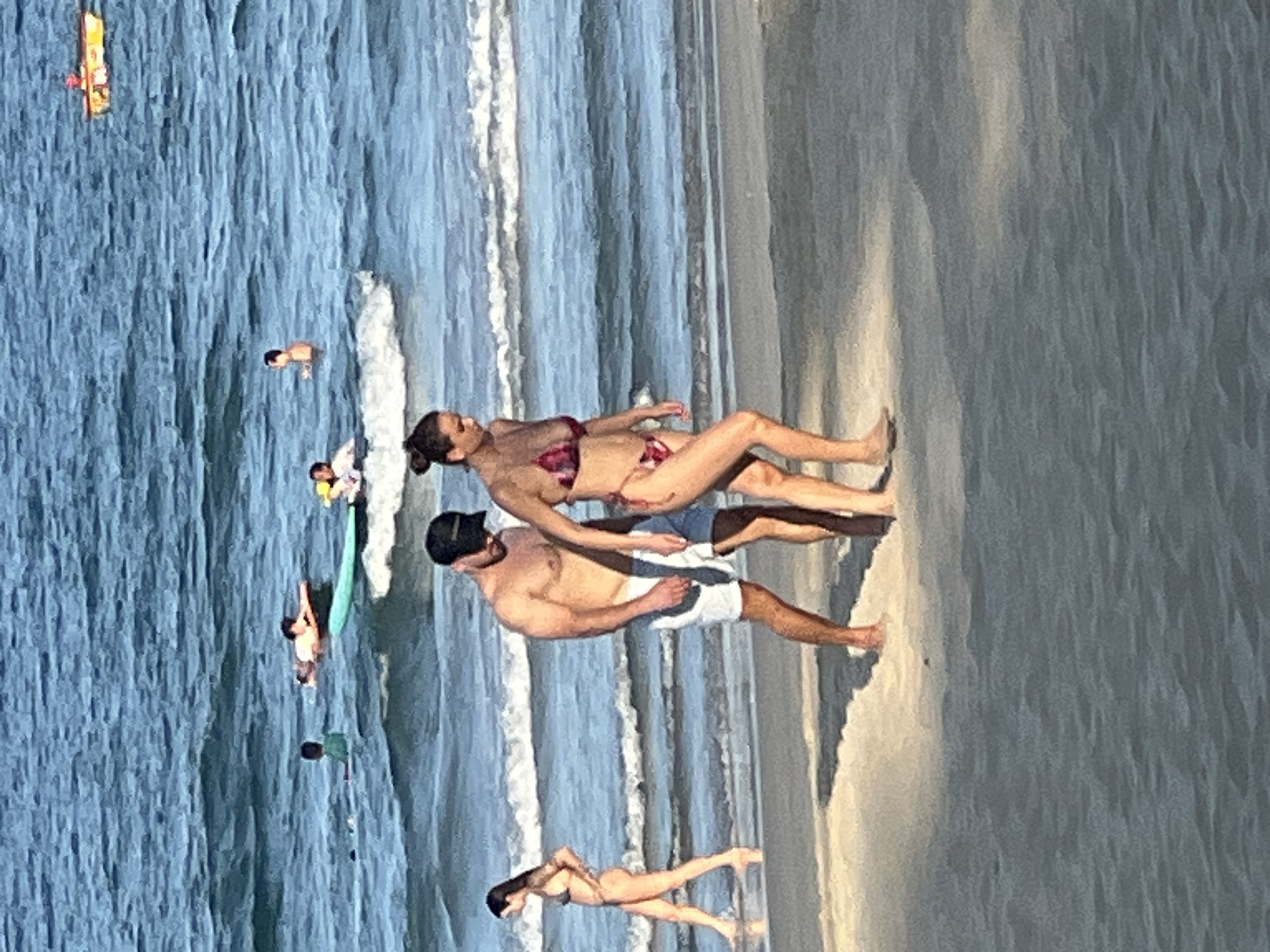 The image captures a daytime scene at a beach in Da Nang, Vietnam. In the foreground, a couple walks along the wet sand near the shoreline. The man is wearing white shorts and a black baseball cap, while the woman is in a patterned bikini. To the left, a woman in a bikini walks away from the camera. In the background, several individuals are visible in the ocean. Some are swimming or wading, while others are on surfboards. A yellow boat with a person is also present further out at sea. The water is blue and wavy, with white foam from the breaking waves. The beach appears sandy and the sun is casting shadows on the sand.