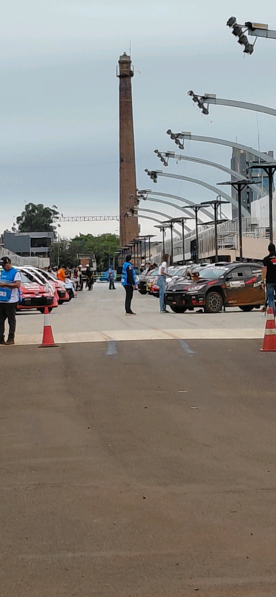 The image shows a street scene in Encarnación, Paraguay, possibly at a motorsport event. In the foreground, a tarmac surface is visible with some markings. Several rally cars, predominantly black and red, are lined up. A tall, brick industrial chimney stands prominently in the background. Overhead, a series of arched structures with attached lighting fixtures are visible. Several people are present, some appearing to be participants or staff in vests, while others are spectators. The sky is overcast.