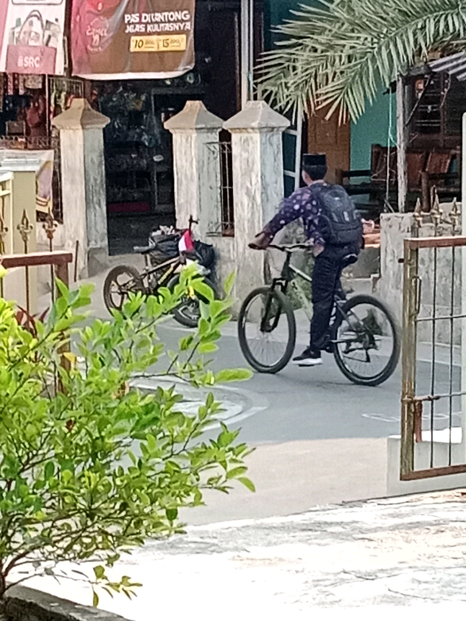 The image depicts a street scene in Waru, Indonesia. In the foreground, a person wearing a black cap and a patterned purple shirt with a backpack is riding a bicycle. The bicycle is black with silver accents and has front and rear suspension. The person is wearing dark pants and white sneakers. To the left of the cyclist, a second bicycle, a BMX type with a red and white element on the handlebars, is leaning against a wall. In the background, there is a building with open doorways and windows, adorned with banners, one of which displays Indonesian text and numbers "10" and "15". A palm frond is visible overhead. The street is paved, and there are decorative concrete pillars and a metal fence. Lush green foliage is present in the lower left portion of the image. The overall impression is that of a casual outdoor scene in a populated area.