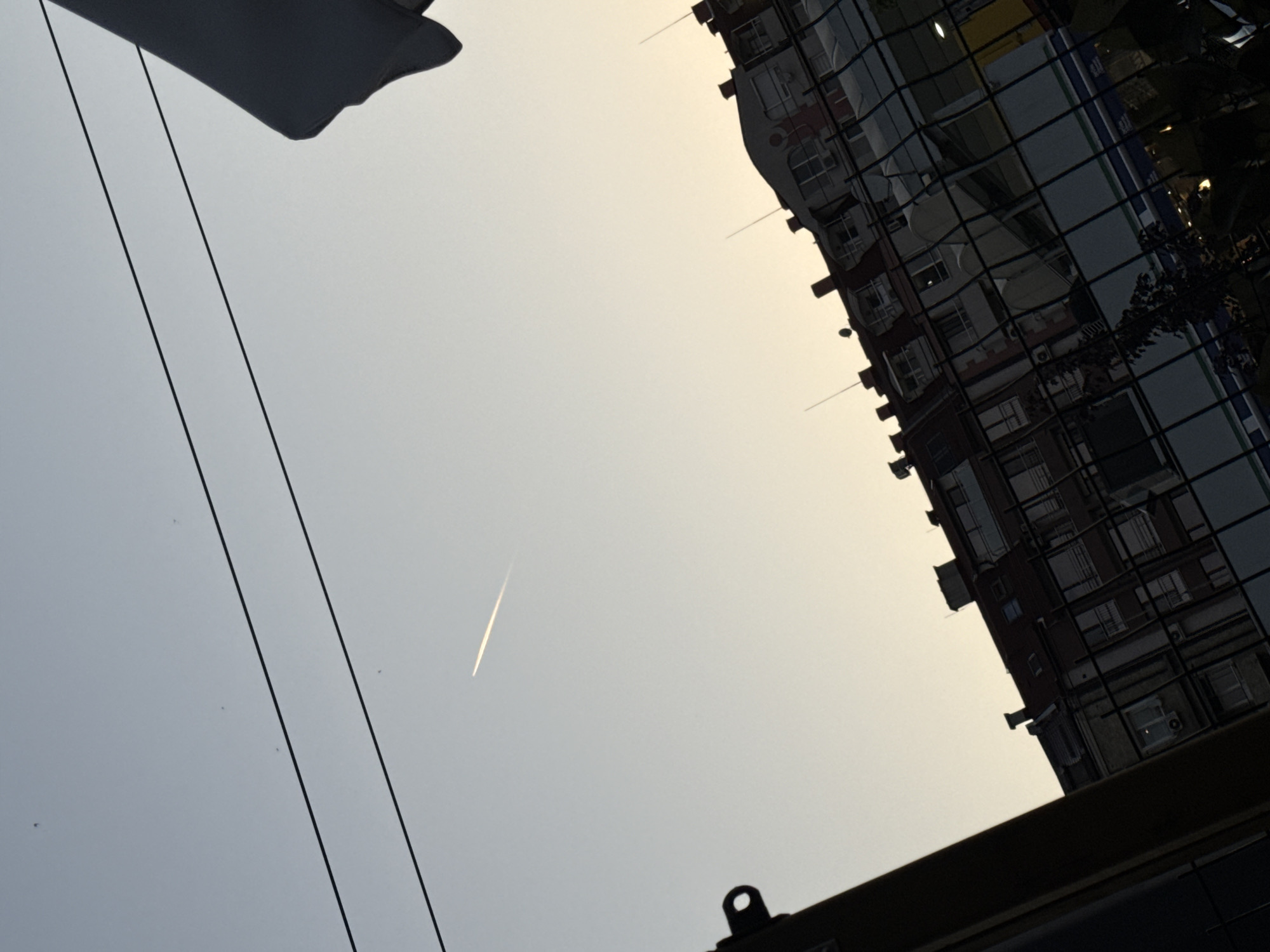 This is a low-angle, tilted shot of a building reflected in glass, with a bright, hazy sky above and some overhead cables. The building's facade appears to be made of glass panels, reflecting other buildings with windows and some greenery. The sky is a light, overcast color, suggesting it might be dawn or dusk, or a generally hazy day. A faint contrail from an airplane is visible in the sky. In the foreground, a dark, angular object, possibly a piece of furniture or part of a structure, is visible at the bottom, partially obscuring the view. The image has a slightly abstract quality due to the angle and reflections. The setting appears to be urban, with the building and overhead cables being characteristic of a city environment. There are no people visible in the frame. The overall mood is somewhat muted and perhaps a little melancholic due to the overcast sky and the starkness of the architectural elements.