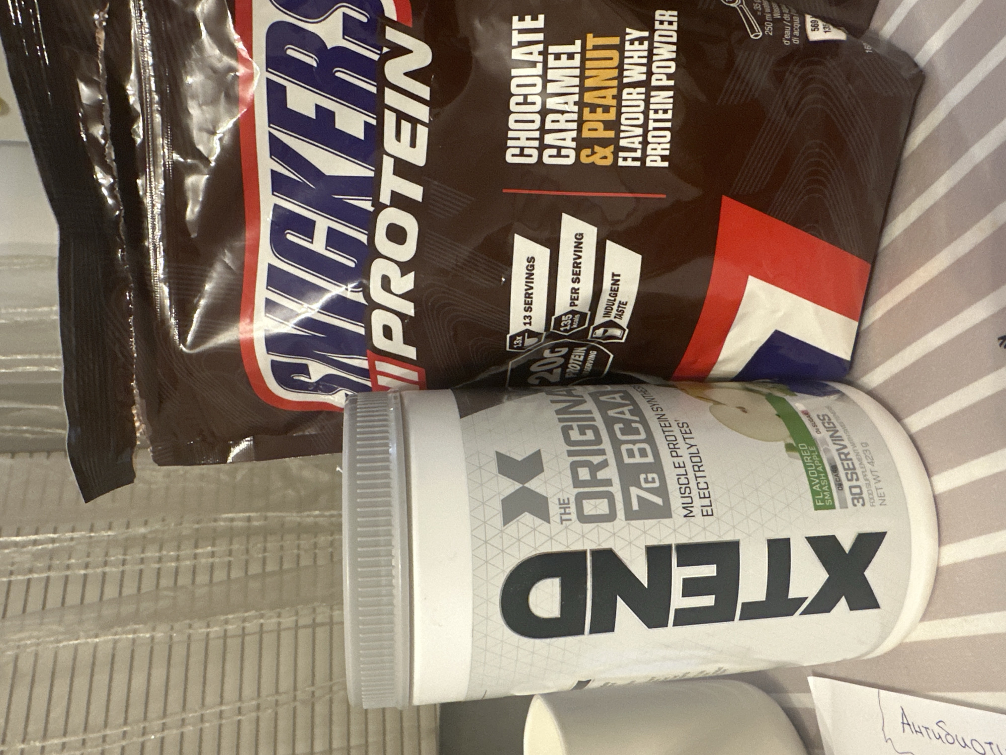 A still, eye-level indoor shot shows two containers of protein powder. A package of Snickers protein powder, labeled as chocolate, caramel, and peanut flavored, is stacked behind a container of XTEND BCAA. The XTEND container is white with a grey patterned background and black lettering. The Snickers package is brown with red and white lettering, and the words "Snickers" and "Protein" are prominently displayed. A portion of a handwritten note in Cyrillic, possibly "Antibiotics", is visible at the lower right. The background is out of focus. The location, Sofia, Bulgaria, is not discernible from the image.