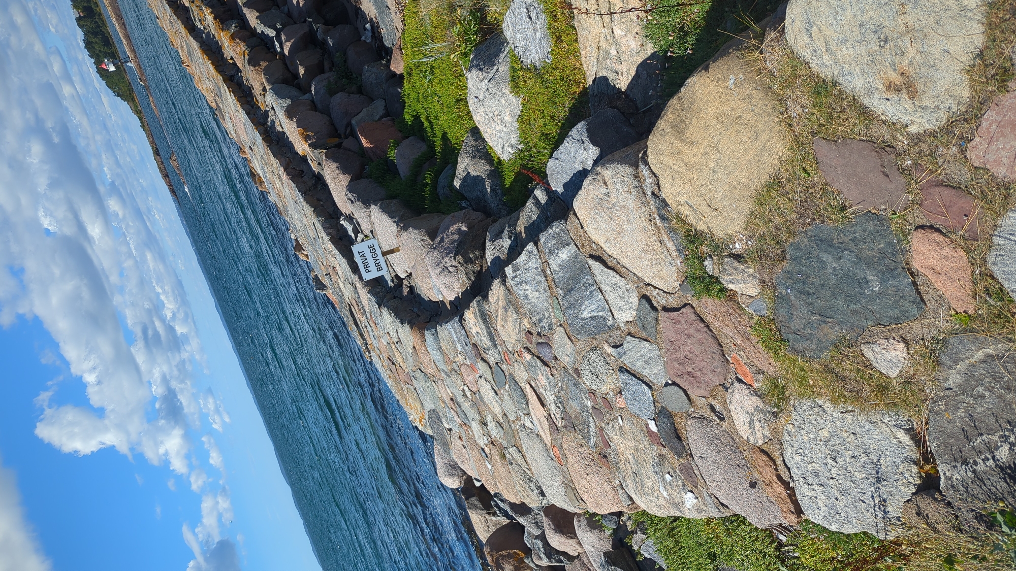 The image shows a stone-lined seawall and a body of water under a partly cloudy sky. A sign on the seawall reads "PRIVAT BRYGGE," which translates to "PRIVATE JETTY." The seawall is made of various sizes and colors of stones, with grass growing in between some of them. The water is a deep blue and appears calm with small ripples. In the distance, a small lighthouse or beacon is visible on a rocky outcrop. The overall scene suggests a coastal area in Larkollen, Norway, characterized by its rocky shores and maritime elements.