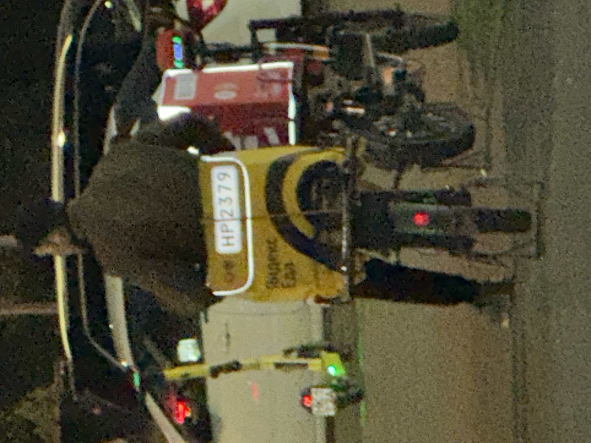 The image shows a delivery person on a motorized scooter with a cargo box. The delivery person is wearing a dark jacket. The scooter has a yellow cargo box with Russian text that appears to be for a food delivery service. A license plate with "HP2379" is visible on the box. The scooter has red and green lights. In the background, a white car is partially visible, and other scooters are also present. The scene appears to be on a street at night in Podolsk, Russia.