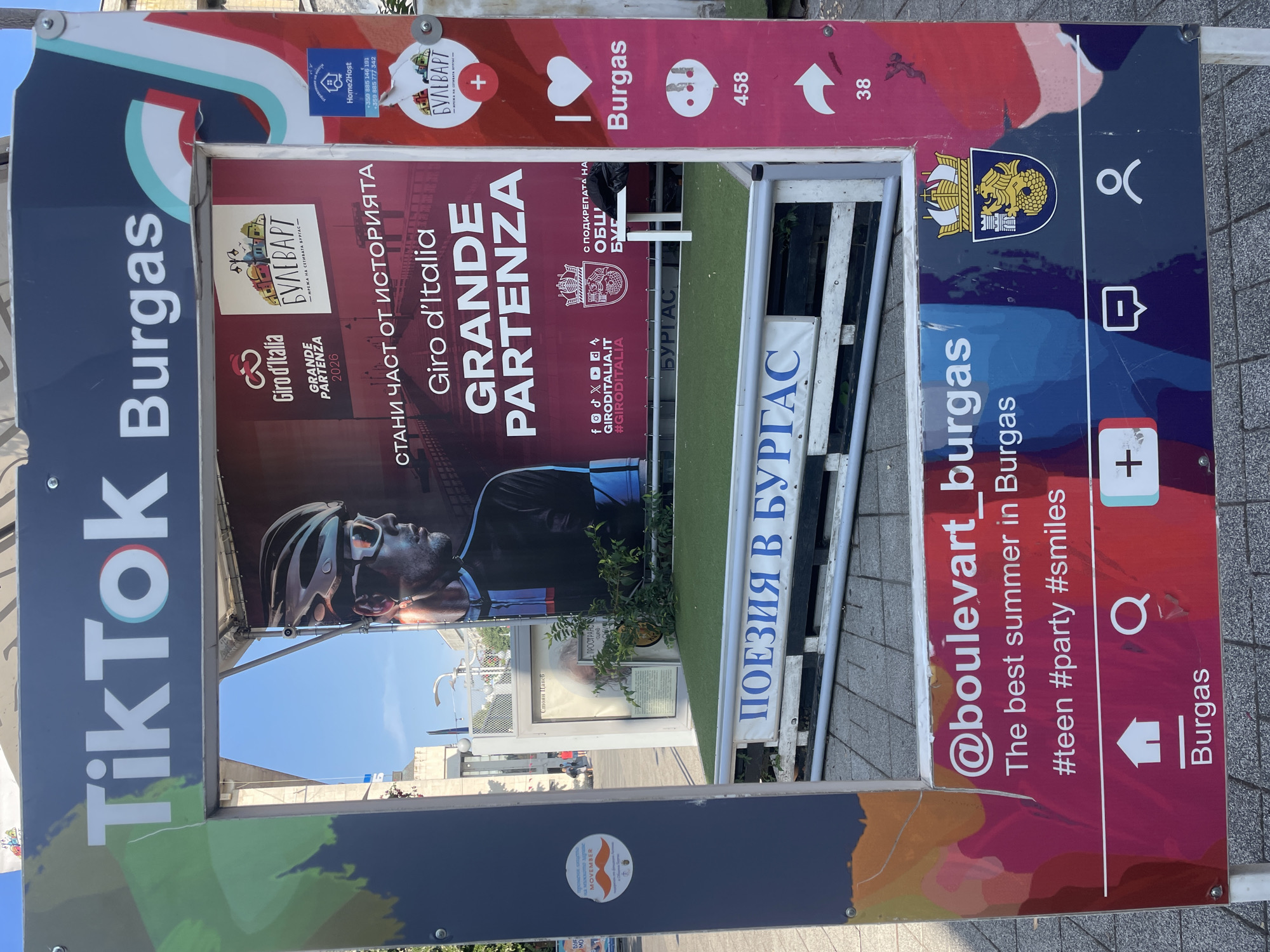 The image shows a large, temporary, Instagram-style photo booth frame set up outdoors, likely in a public square or promenade in Burgas, Bulgaria. The frame itself is decorated with vibrant colors and graphics, featuring the TikTok logo prominently at the top, followed by "Burgas." The background visible through the frame includes a poster advertising the Giro d'Italia Grande Partenza 2026 event, which is stated to be in Burgas. Another poster features a cyclist's profile.
The photo booth structure is built on a raised platform covered with artificial grass, with a banner displaying "ПОЕЗИЯ В БУРГАС" (Poetry in Burgas) in large blue letters. A small white table is visible behind this banner.
On the right side of the frame, there are social media-style icons and text. A pink panel displays a heart icon and the word "Burgas," followed by a speech bubble icon with "458" below it, and an arrow icon with "38" below it.
On the bottom part of the frame, there's a section designed to mimic a social media post. It includes the username "@boulevart_burgas," followed by the text "The best summer in Burgas" and hashtags like "#teen #party #smiles." Below this are icons representing a house, a magnifying glass, a plus sign, a speech bubble, and a person silhouette. The word "Burgas" is also displayed below the house icon. The coat of arms of Burgas is visible on the right side of this section.
The overall scene suggests a promotional event or attraction designed to engage the public, likely related to tourism or a sporting event. The setting appears to be a sunny day, with some buildings and sky visible in the background. There are no people clearly visible in the foreground or interacting with the photo booth, though a few individuals can be faintly seen in the distant background.
