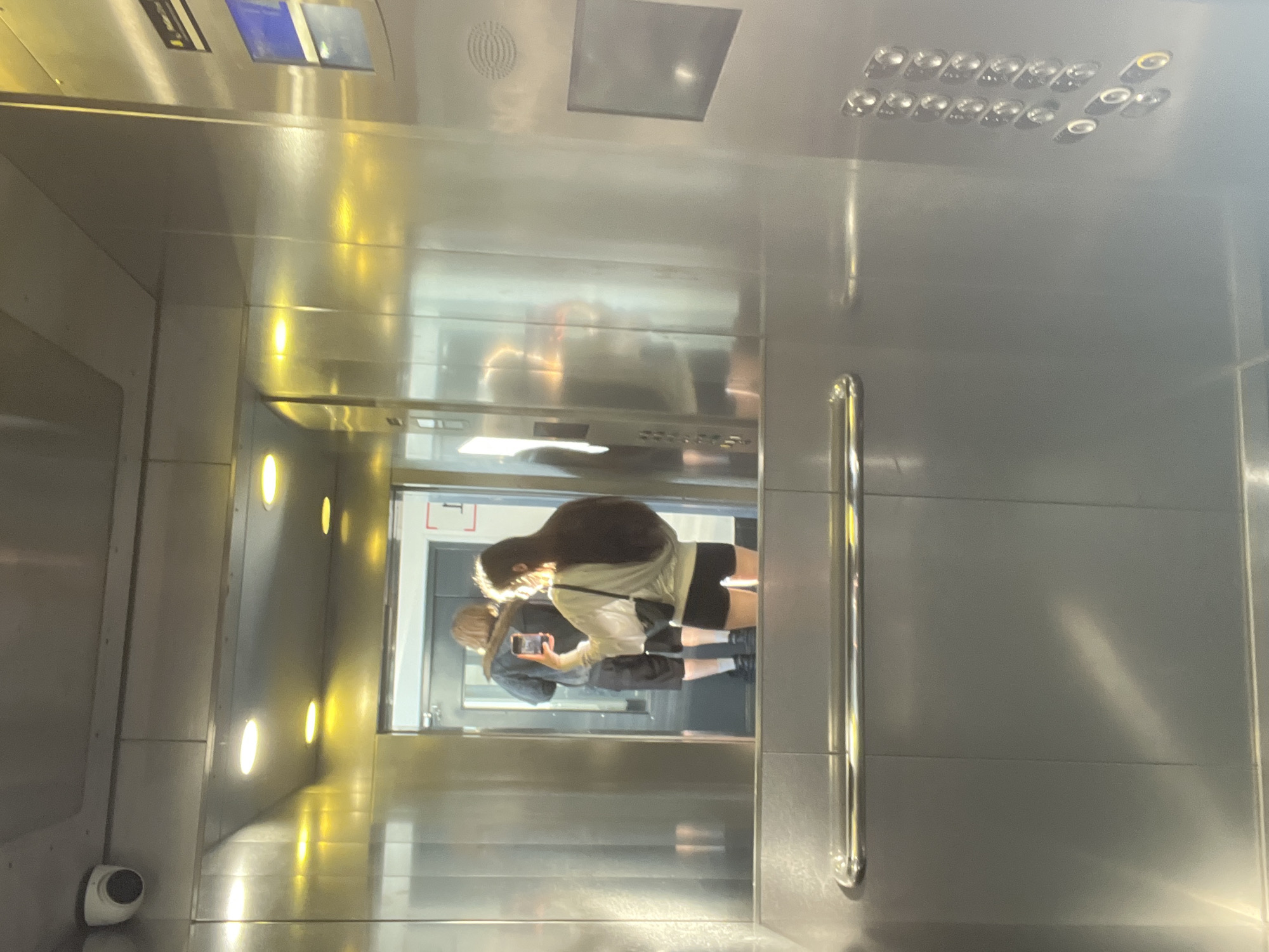 The image captures the interior of a modern elevator cabin with polished metallic walls, which function as reflective surfaces. Two individuals are visible in the reflection on the rear wall of the elevator. In the foreground, a female with long, dark hair, wearing a light-colored long-sleeved top and dark shorts, holds a smartphone horizontally, appearing to take a photograph or video. She has a small black bag slung across her body. Behind her, a male individual with lighter hair is seen from the back, wearing a dark jacket or shirt and dark shorts. Both subjects are positioned facing the open elevator doors.
The elevator cabin is illuminated by multiple recessed circular lights in the ceiling, casting a warm yellow glow. A white security camera (CCTV) is mounted in the upper left corner of the ceiling. A horizontal chrome handrail extends across the lower section of the reflective wall. On the right-hand wall, a control panel features two vertical columns of numerous oval-shaped buttons, with two larger circular controls positioned below them. Above the buttons, there is a small digital display and a speaker grille. A black sign with white text is partially visible in the upper right corner of the elevator, with discernable characters suggesting "Peterburg" or "Petersburg." The open elevator doors reveal a well-lit hallway or landing beyond.
The scene is located in the city of Saint Petersburg, Russia.
