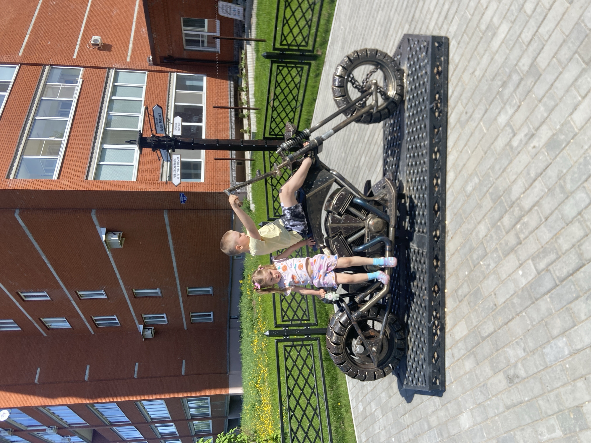 Two children, a boy and a girl, are interacting with a motorcycle sculpture. The boy is sitting in the driver's seat, while the girl is positioned on the motorcycle's frame. The sculpture rests on a black metal platform set on a light gray brick pavement. Behind the sculpture, a green lawn is bordered by a black wrought-iron fence. In the background, a multi-story red brick building stands. A signpost with multiple directional signs points towards various locations, including Kapra Naritsa and the Chaikovsky Park of Culture and Rest. The scene is located in the city of Chaikovsky, Russia.