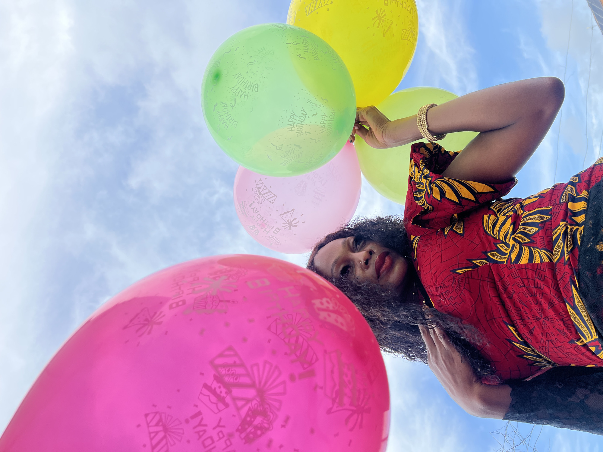 An outdoor, low-angle shot captures a woman holding a cluster of colorful "Happy Birthday" balloons against a bright, cloudy sky in Igboefon, Nigeria. The woman, positioned in the upper right of the frame, has dark, curly hair and looks directly at the camera with a neutral expression. She is wearing a vibrant red and yellow patterned top, possibly made from an African print fabric, with black lace visible on her arm. Her right arm is adorned with a gold-colored beaded bracelet as she holds the balloon strings. A subtle nose piercing is visible on her face, and her lips are colored with red lipstick.
The foreground is dominated by a large, bright pink balloon, profusely decorated with repeated "HAPPY BIRTHDAY" text and various birthday-themed motifs like cakes, presents, and party hats. Behind it, a cluster of green, yellow, and lighter pink balloons also display similar "HAPPY BIRTHDAY" greetings and celebratory graphics. The background features a soft blue sky with scattered white, diffused clouds, indicating a daytime setting. The overall scene conveys a celebratory atmosphere, likely a birthday event.