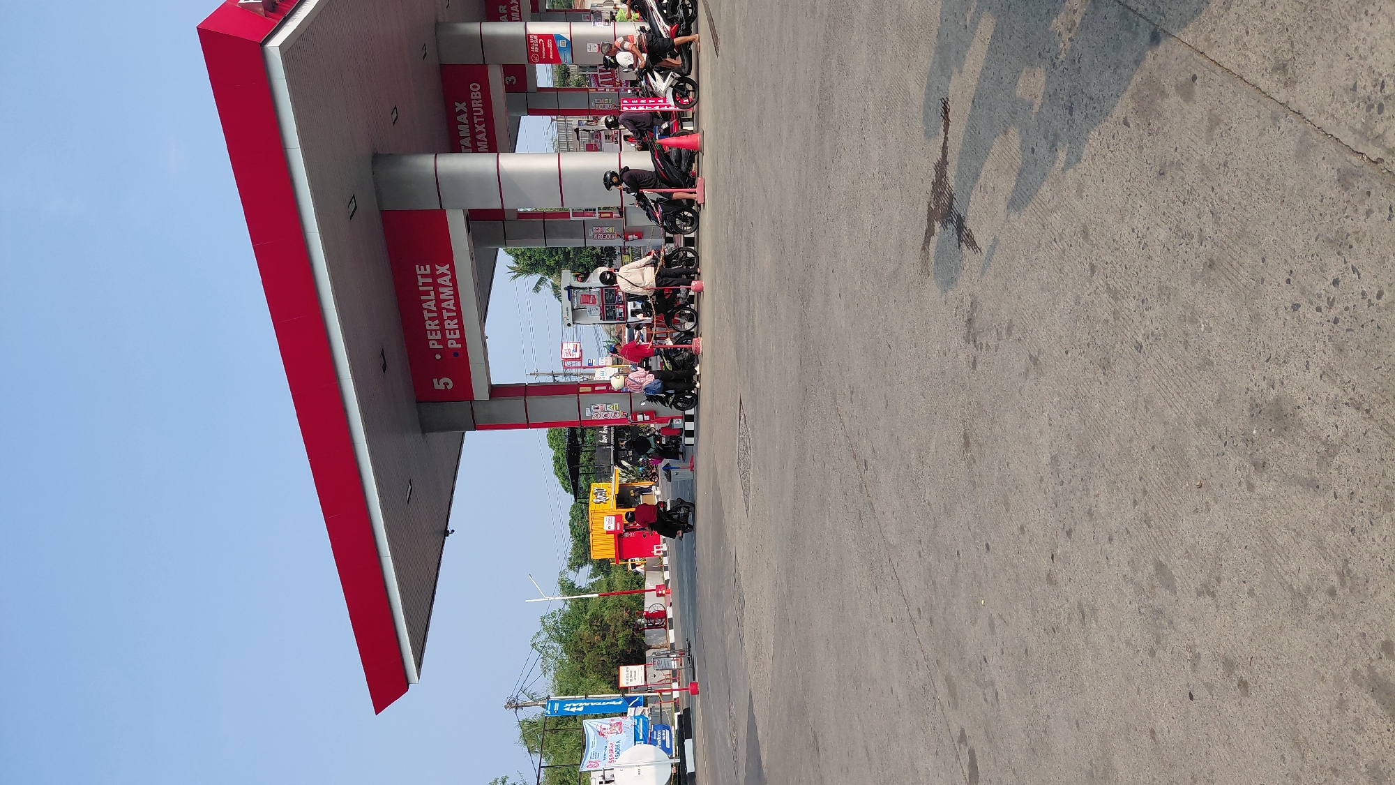 This image depicts a Pertamina gas station located in Karanglewas, Indonesia. The scene is characterized by a bright red and white canopy over multiple gas pumps. Several individuals on motorcycles are present, either refueling or waiting to refuel. One individual is seen wearing a red shirt and black helmet on a motorcycle in the foreground. Another individual, also on a motorcycle, is wearing a light-colored shirt and a white helmet. A third individual, dressed in a pink shirt and white helmet, stands next to a motorcycle. The ground is paved concrete, with some scattered dark stains. In the background, there are trees, other small buildings, and banners. The sky is clear and blue. The overall atmosphere suggests a typical, busy day at a gas station.