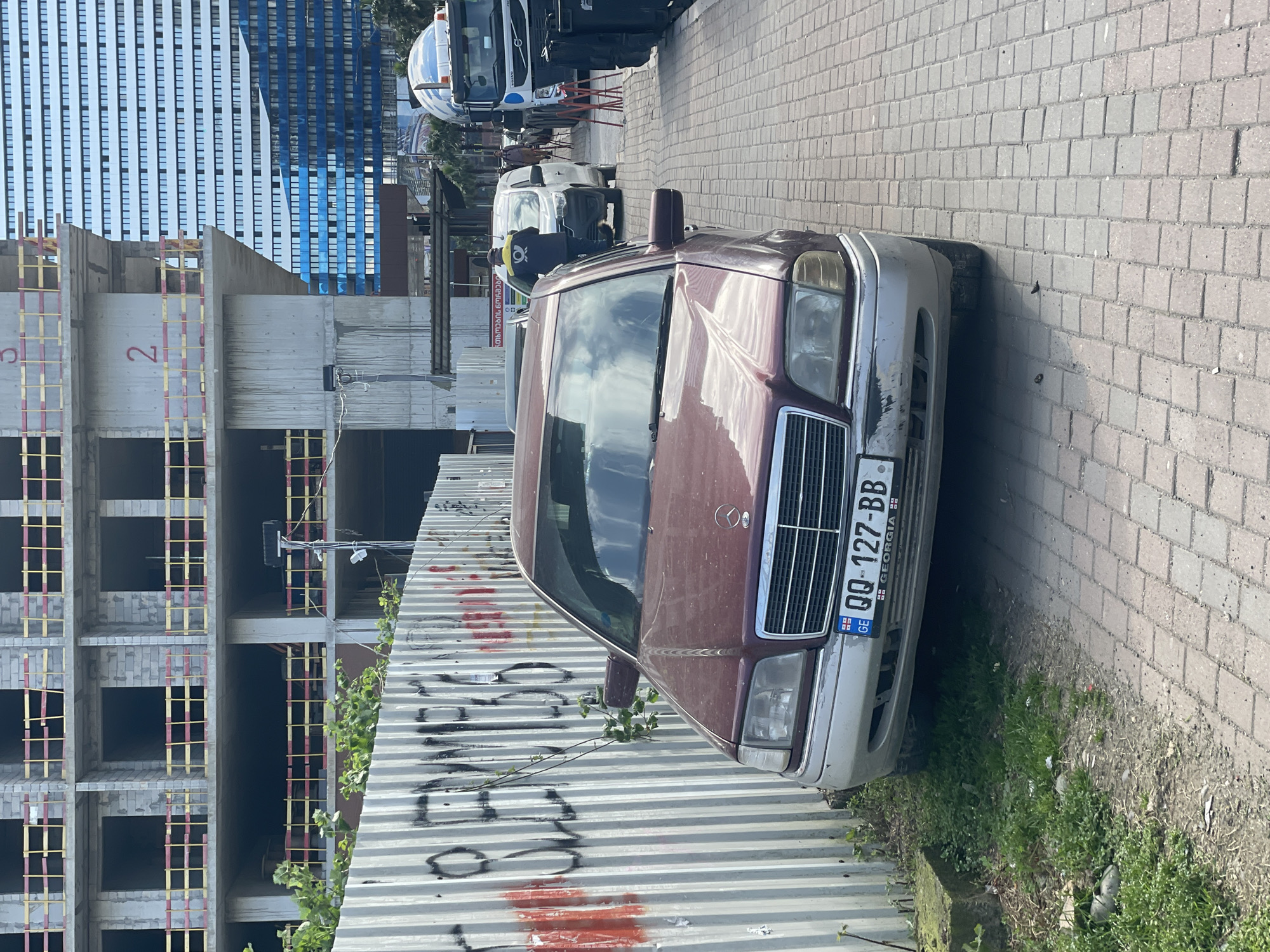The image shows a maroon Mercedes-Benz parked on a brick sidewalk next to a corrugated metal fence with graffiti. The license plate reads "QQ-127-BB" and indicates the car is registered in Georgia. Behind the fence, a building under construction with scaffolding is visible. In the background, tall buildings with a blue and white facade can be seen. The location is likely Batumi, Georgia, given the Georgian license plate and the style of architecture. Other vehicles and people can be seen further down the street. The scene appears to be a typical urban environment with construction activity and parked vehicles.