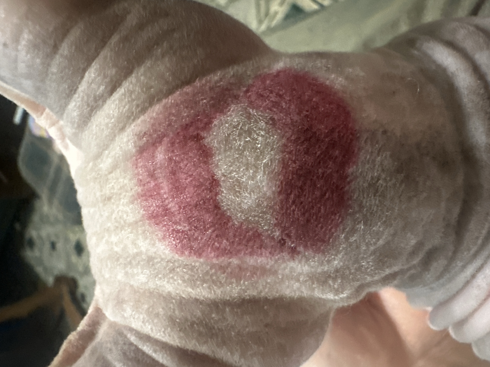 The image shows the back of a hairless cat's head with a red lipstick mark on it. The mark is roughly circular with a lighter center, resembling a stylized heart or a kiss. The cat's skin is wrinkled, and it is wearing a light-colored garment around its neck. The background is out of focus, with what appears to be a patterned rug or blanket and a dark object that might be furniture. There is no indication of the location being in Togliatti, Russia, or any other specific city. The image is a close-up of the cat's head and does not provide any contextual information about the environment or its surroundings.