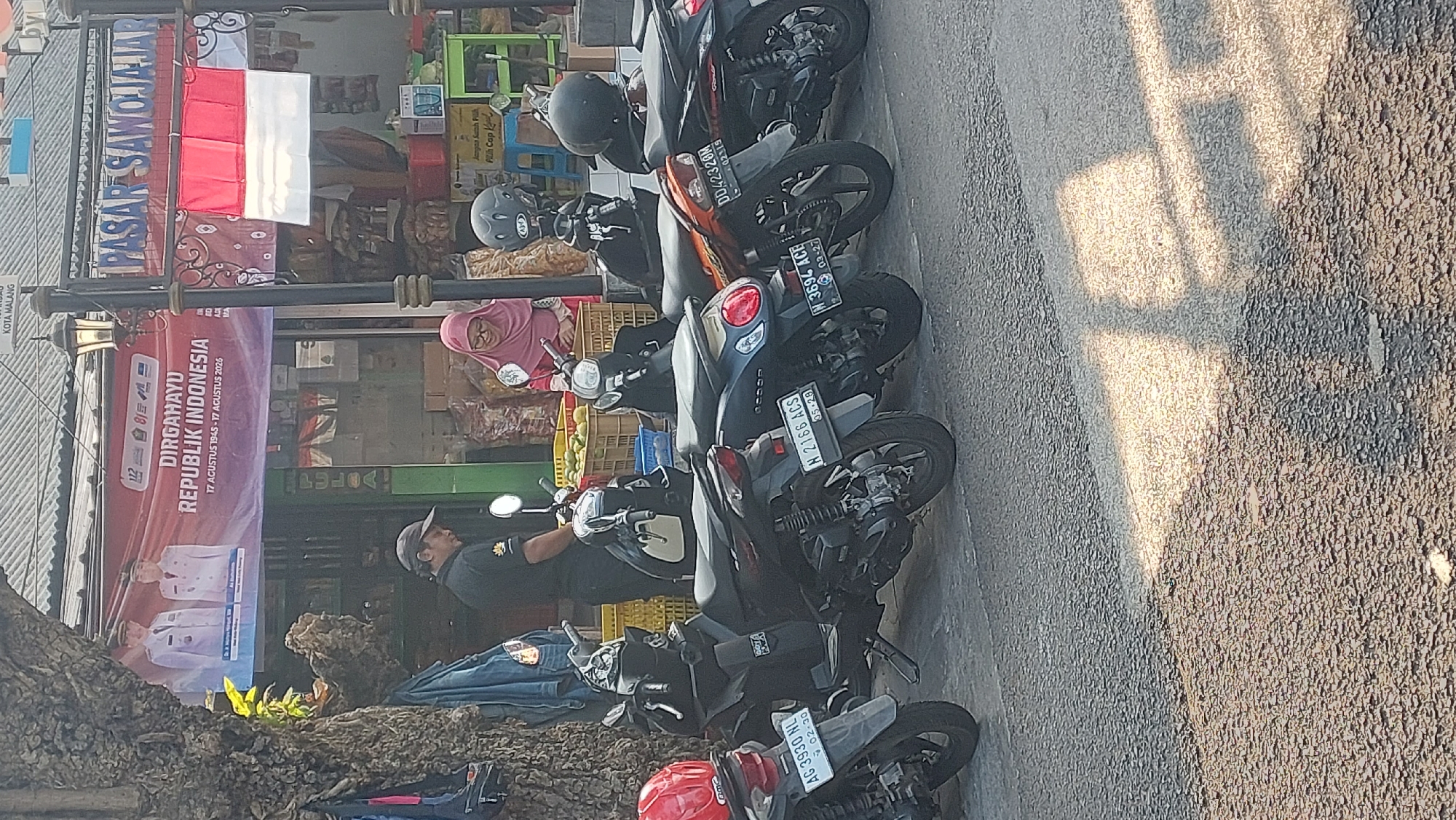 A row of parked motorcycles is visible on a paved street in front of shops in the city of Malang, Indonesia. In the background, a banner reads "DIRGAHAYU REPUBLIK INDONESIA" and features images of two men in formal attire. A flag of Indonesia is displayed above a sign that says "PASAR SAWOJAJAR". Two individuals, one wearing a pink hijab and the other a black shirt and cap, are interacting near the shops. The scene is illuminated by sunlight, casting shadows on the ground.