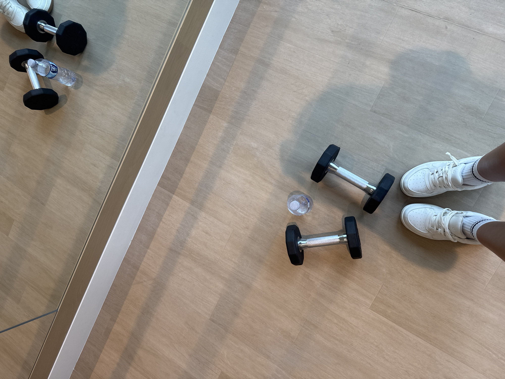 A first-person perspective shot shows a person's white sneakers and ankles standing on a light brown, wood-grain floor. In front of the person, two hexagonal dumbbells with black weights and silver handles are placed on the floor. Between the dumbbells, a partially filled clear plastic water bottle with a white cap is situated. A mirror runs along the top of the image, reflecting two more dumbbells and a water bottle. The scene appears to be indoors, likely in a gym or home exercise space.
