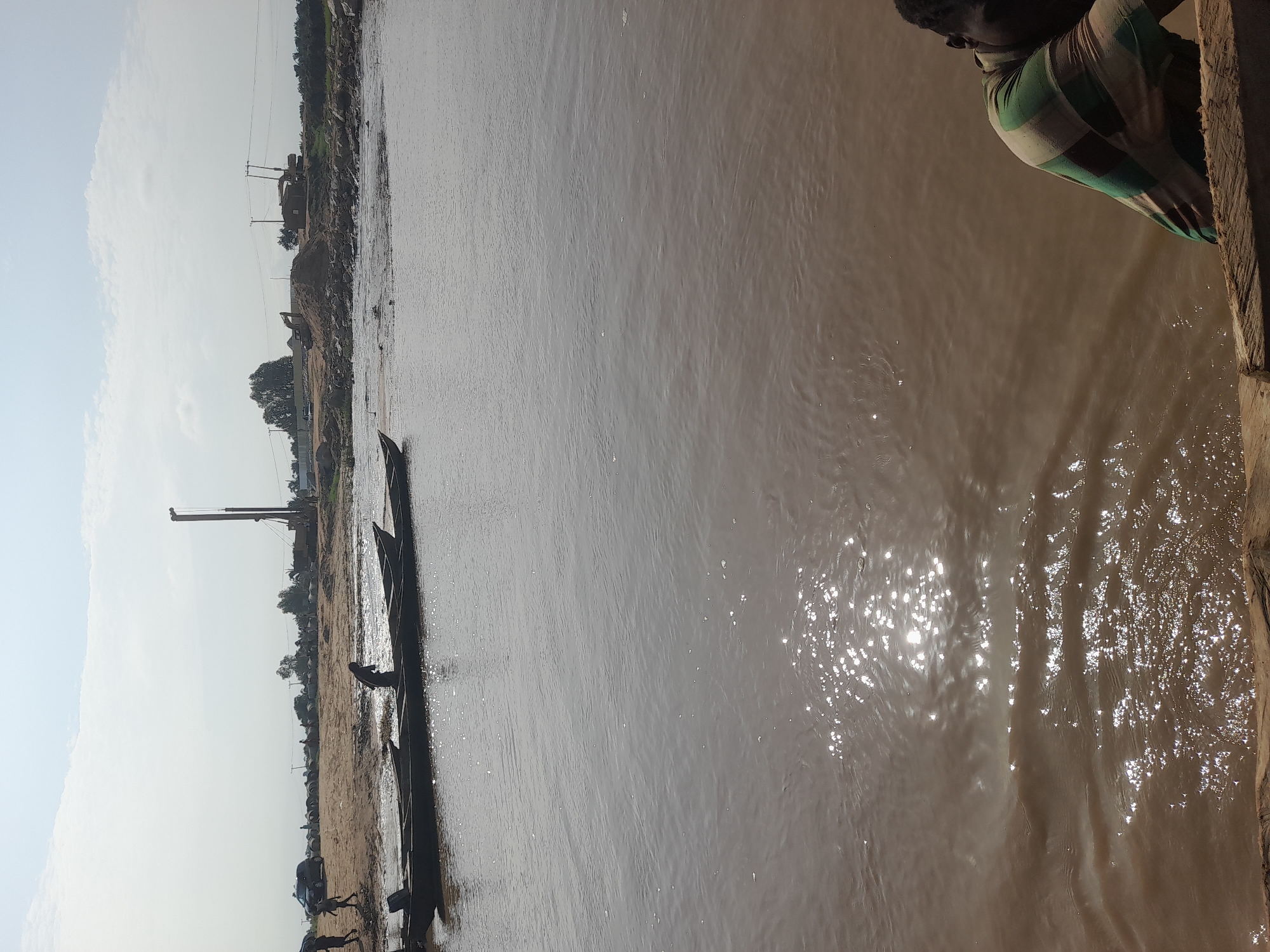 In Nemnei, Nigeria, a wide, muddy river occupies the foreground, reflecting the hazy sky and a bright sun. Several long, narrow wooden boats are moored along the sandy bank on the left side of the image. One person is visible in one of the boats. Further inland, construction equipment, including a large drilling rig, excavators, and piles of pipes, suggests ongoing development. Silhouetted figures of people are scattered across the sandy area. Power lines stretch across the background, with trees and buildings visible in the distance. In the lower right corner, a person wearing a horizontally striped shirt is partially visible, leaning on a wooden structure.