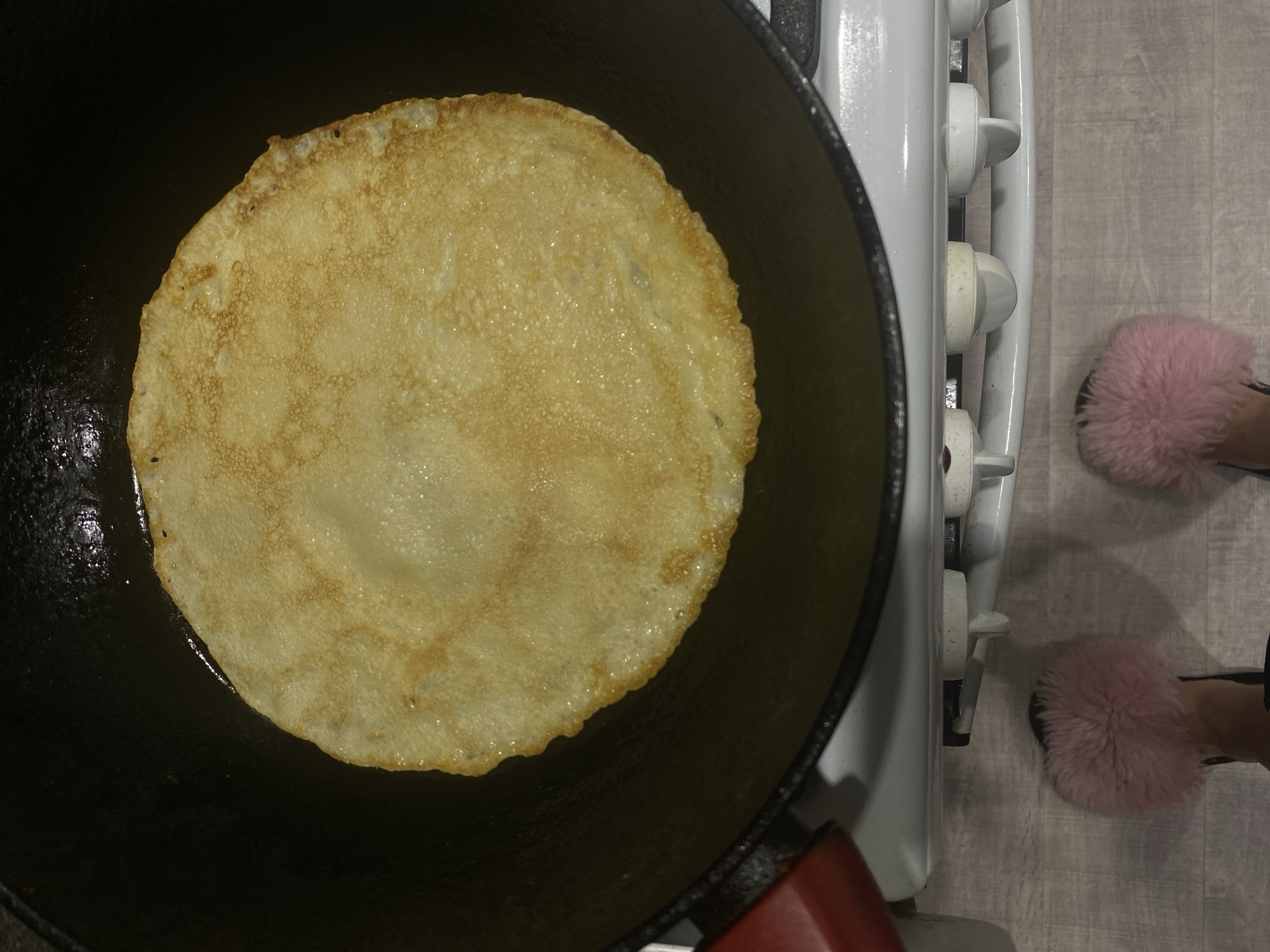 A person is observed from an overhead perspective, standing in a domestic kitchen. The primary subject is a circular, light golden-brown pancake or crepe cooking in a dark frying pan, which occupies the upper two-thirds of the image. The pancake exhibits slight browning and a textured surface. The pan, likely cast iron, has a red handle visible on the lower left side. It rests on a white gas stove, the control knobs of which are partially visible below the pan. Below the stove, a person's feet are visible, wearing pink, fluffy slide slippers. The floor beneath the feet is a light-colored surface, possibly laminate flooring. The image provides no specific details to determine a precise location within the city of Sochi, Russia.