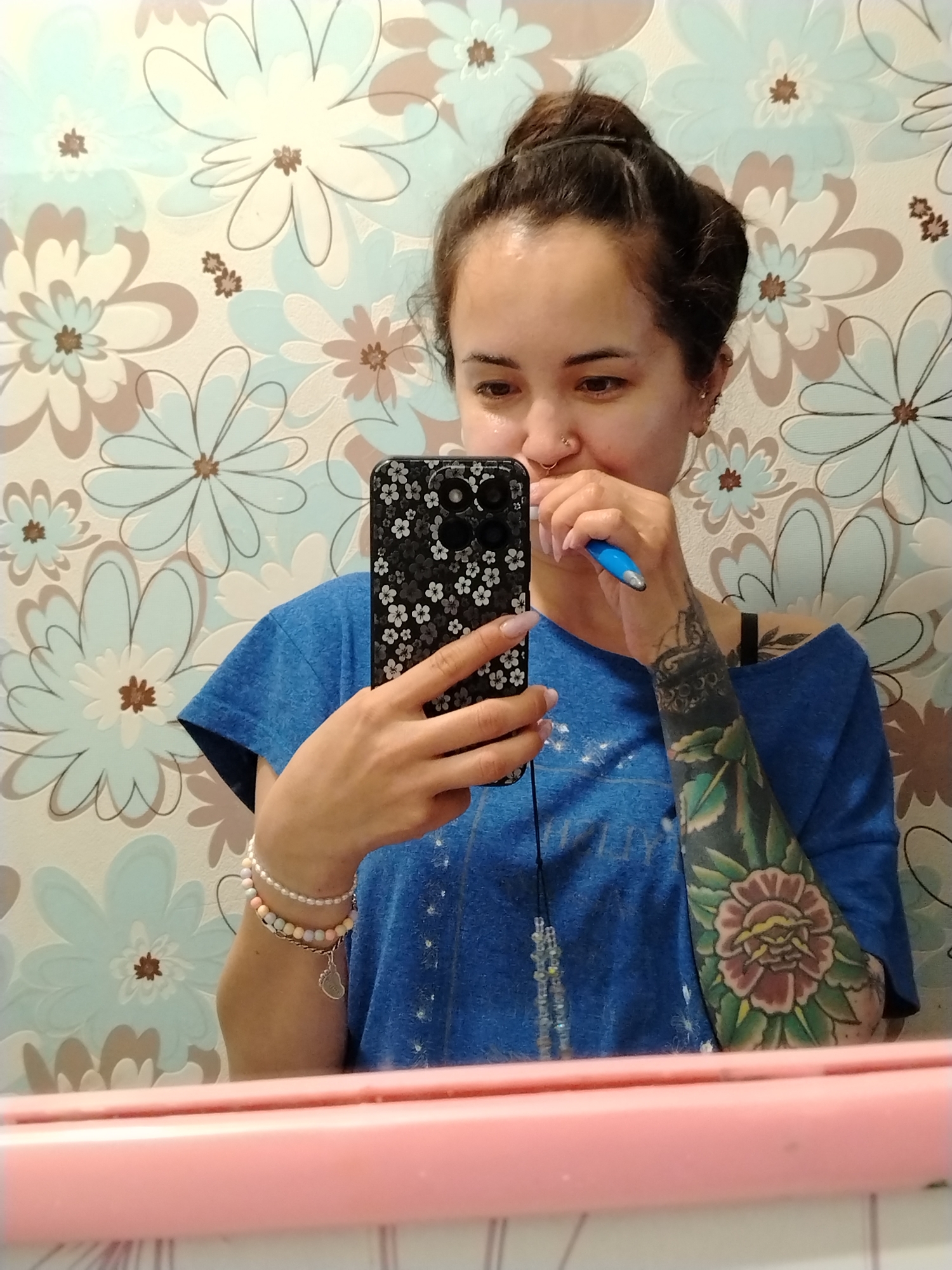 A woman is visible in a selfie taken in a bathroom, brushing her teeth. The background includes a light blue and beige floral wallpaper. She is wearing a blue t-shirt and has a full-sleeve tattoo on her left arm. She also has bracelets on her left wrist and a nose ring. The location is a bathroom in an Unknown city.