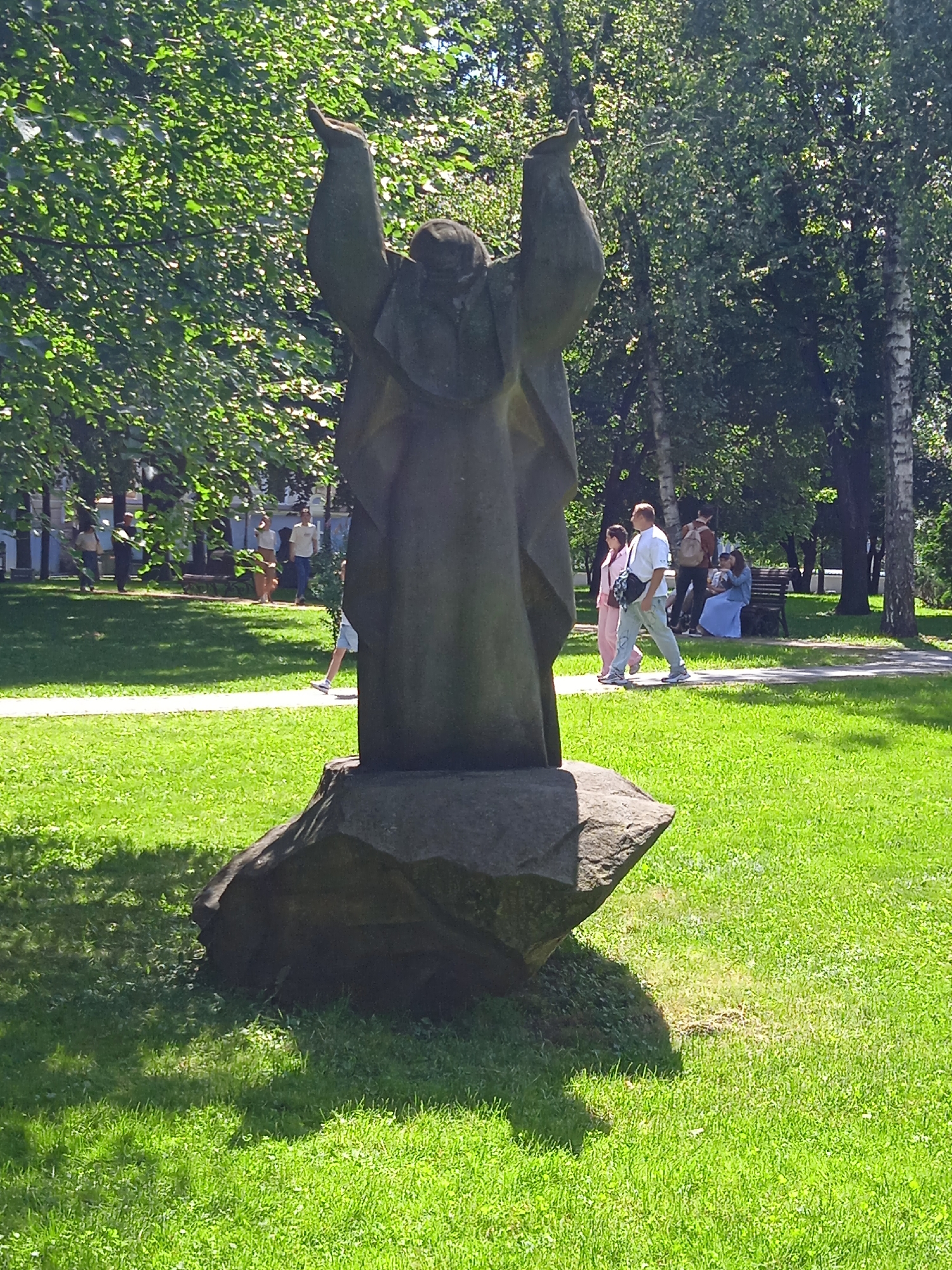 The image shows a stone sculpture of a robed figure with arms raised, situated on a large rock in a grassy park. The sculpture is dark grey and abstract in style. In the background, several people are walking on a paved path. To the left, a few individuals are seen near some trees. To the right, a couple is walking, and further back, a woman in a blue dress is sitting on a bench with a child. The environment is a sunlit park with abundant green grass and trees. The sculpture is located in Shevchenko Park, Kyiv, Ukraine, specifically near the entrance to the park.