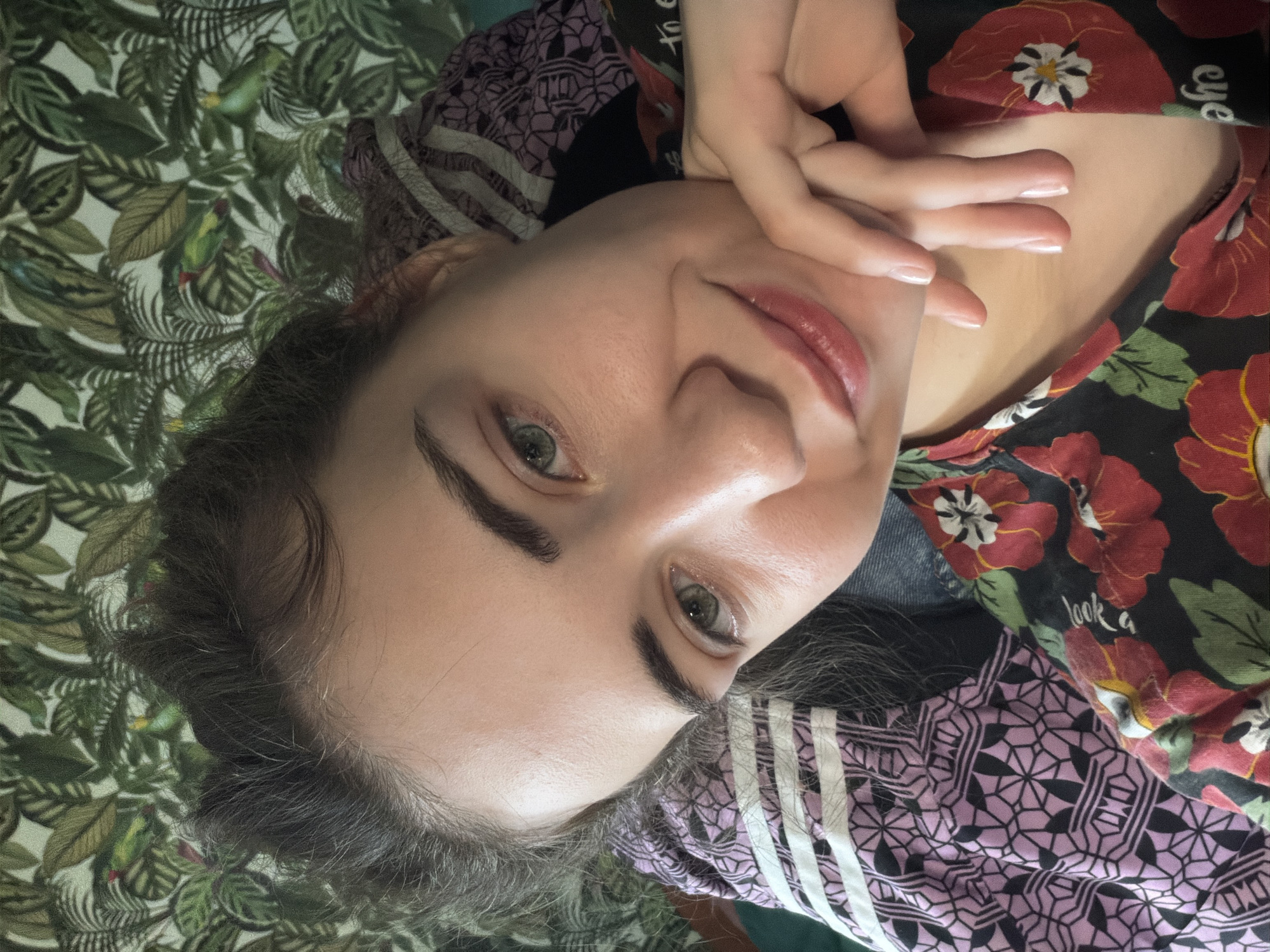A young woman with light skin, dark hair pulled back, and green eyes is photographed from the chest up. She is leaning on her hand, with her chin resting on her knuckles. She is wearing a black t-shirt with a floral print featuring red poppies and green leaves. Behind her is a wall with a tropical-themed wallpaper depicting palm leaves and two green parrots. The woman appears to be indoors, possibly in a bedroom or living room. There is no information in the image to suggest a specific location in Zavodoukovsk, Russia.