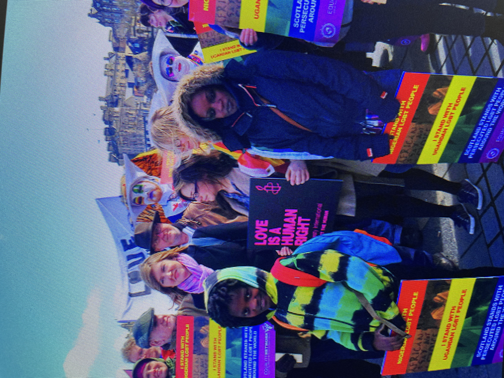 A group of people are holding signs that support LGBTQ+ rights. One sign reads "Love is a Human Right." Several signs mention specific countries, including "Uganda LGBT People" and "Nigeria LGBT People." Other signs state "Scotland Stands With Persecuted LGBT People Around the World." The background shows a city scene with buildings in the distance. The photo appears to have been taken during a protest or demonstration. It's daytime and the sky is light. There is no evidence that this image depicts Zlatograd, Bulgaria.