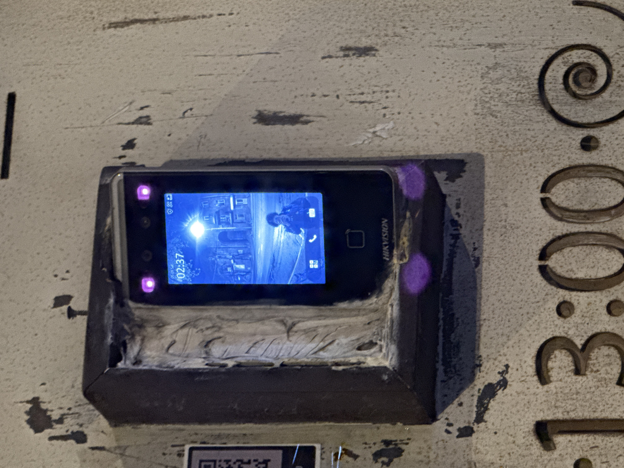 The image shows a Hikvision video intercom device mounted on a textured, light-colored surface. The device's screen displays a video feed, likely from a security camera, showing an exterior scene with buildings and a vehicle. The time on the screen reads "02:37 05.09 Sat". The mounting appears to be custom-made. To the right of the device, carved into the surface, are the numbers "13:00". The location is Yerevan, Armenia based on the reference to "13:00", corresponding to 1:00 PM, a common timestamp reference within the city.