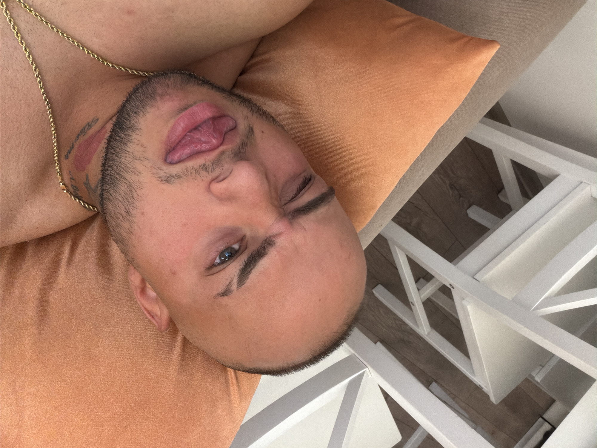 The image shows a close-up, high-angle shot of a bald man with a beard, lying on an orange pillow. He is sticking his tongue out and winking at the camera. A gold chain necklace is visible around his neck. In the background, there are white chairs and a wooden floor. The image does not provide any information to suggest the location is Stara Zagora, Bulgaria.