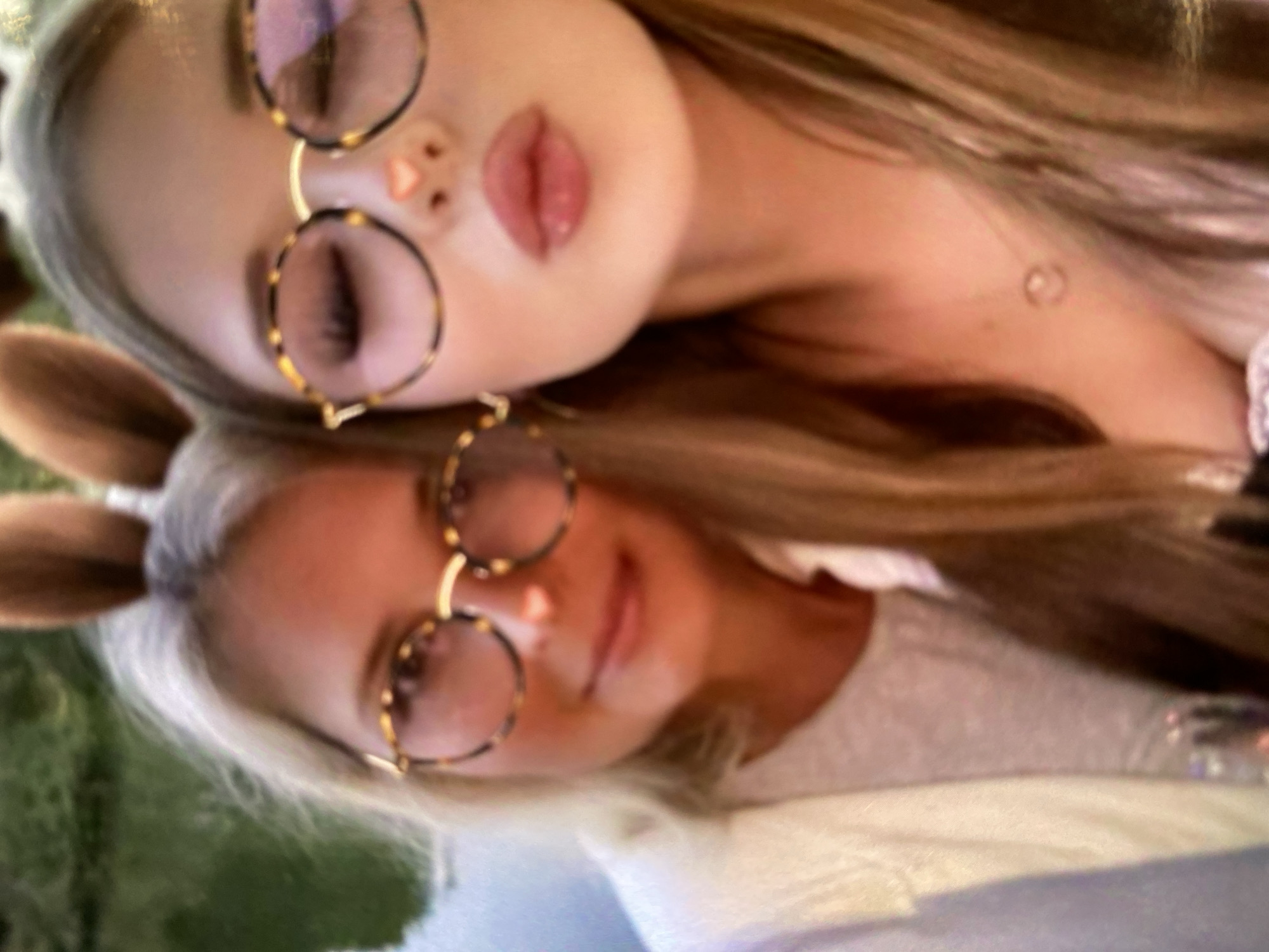 Two women, appearing to be mother and daughter, are posing for a selfie outdoors. They are both wearing large, round, tortoise-shell patterned glasses with pinkish lenses. The woman on the left has blonde hair and is wearing a white cardigan over a light-colored shirt. The woman on the right has blonde hair and is wearing a white top. Both women are wearing fuzzy brown bunny ears. The woman on the right is puckering her lips for the photo, and has a small pink heart shape drawn on her nose to resemble a bunny nose. The background appears to be a body of water and greenery, suggesting a park or waterfront location. There is no indication of this location being in Moscow, Russia.