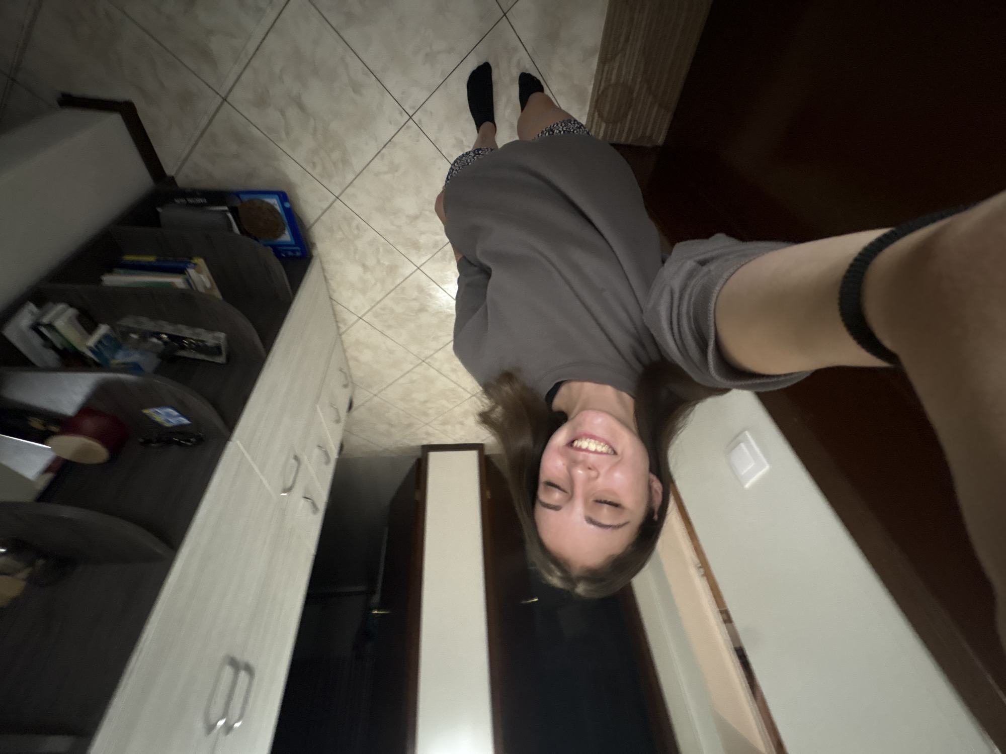 In an indoor setting, a young woman with brown hair, eyes closed, and a wide smile, is taking a selfie. She is wearing a gray long-sleeved shirt and patterned shorts, along with black socks. She appears to be in a room with tiled flooring and cabinetry. The scene is consistent with a residential interior in Bulgarovo, Bulgaria.