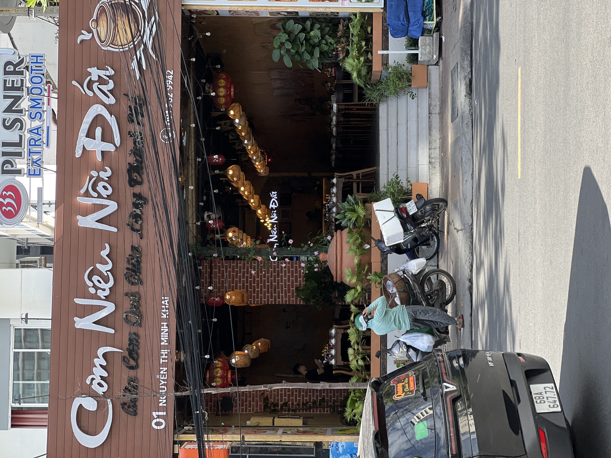 The image shows the exterior of a restaurant called "Cơm Niêu Nồi Đất" located at 01 Nguyễn Thị Minh Khai in Nha Trang, Vietnam. The restaurant has a brown wooden facade with large white cursive lettering spelling out the name. Below the main name, smaller black lettering reads "Cơm Niêu Quê Hiền Long Thương Nhớ".
In front of the restaurant, a black car with license plate "68A 647.72" is parked on the left side of the frame. To the right of the car, a person wearing a light green shirt and dark patterned pants is standing next to a motorcycle, holding a basket filled with items and a plastic bag. Another motorcycle is parked behind them.
The entrance to the restaurant features a set of stairs leading up to a doorway, with plants in planters on either side. Inside, tables are set for dining, and various decorative lanterns hang from the ceiling. Overhead, numerous black electrical wires are strung across the front of the building. In the background, a sign with "333 Pilsner Extra Smooth" is visible. The scene appears to be set during the daytime, with bright sunlight casting shadows.