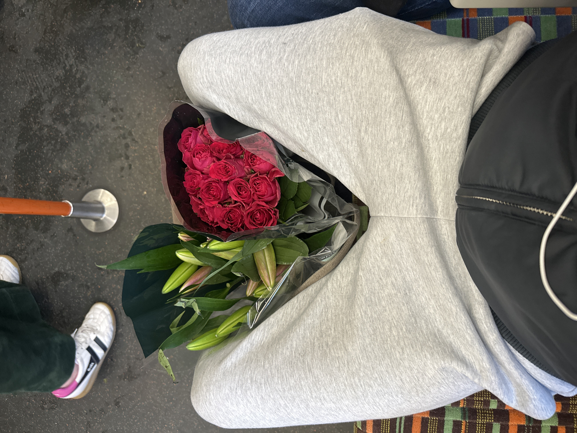 The image depicts an overhead shot of a person sitting on what appears to be public transport, likely the London Underground. The individual is wearing a light grey hoodie and dark jeans. They are carrying a bouquet of red roses and lilies wrapped in clear plastic. To the left, part of the carriage interior is visible, including a handrail and the lower part of a person's leg wearing a green pant and a white Gola sneaker with black stripes. The seating appears to be upholstered with a multicolored, plaid pattern. Overall, the scene conveys a sense of urban life and transport.