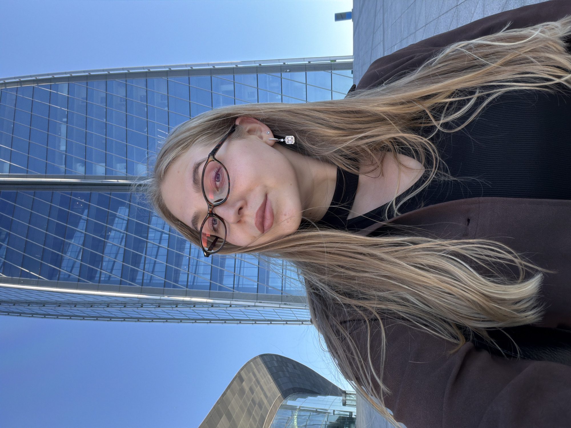 A young woman with long blonde hair and glasses stands outdoors in front of a modern skyscraper with a glass facade, the Lakhta Center in Saint Petersburg, Russia. She is wearing a brown jacket over a black top and a choker necklace, with a square-shaped earring in her left ear. The sky is clear and blue. The building's reflective glass creates geometric patterns in the background.
