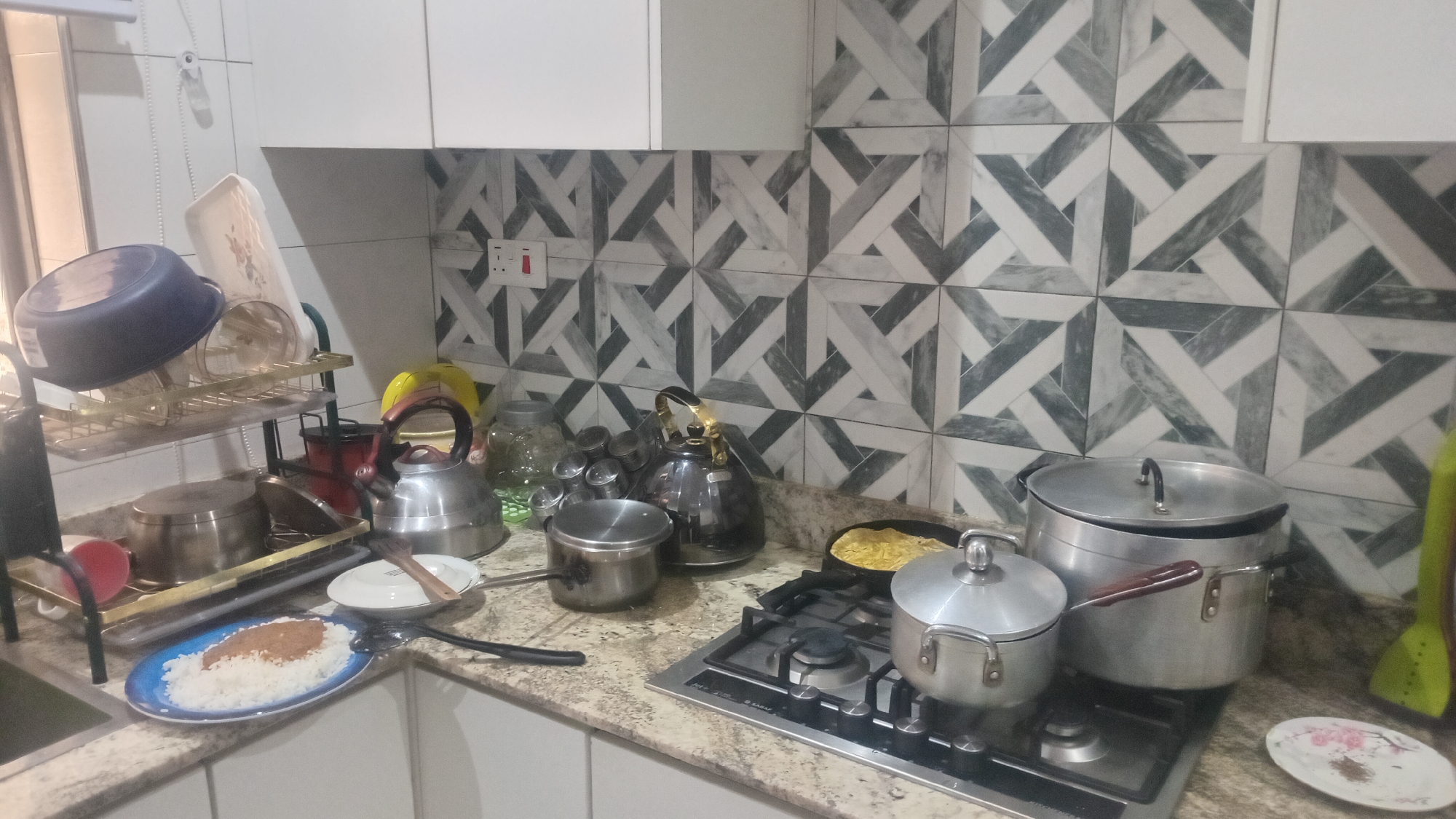 The image shows a messy kitchen counter with various cooking utensils and food items. In the foreground, a blue plate contains a pile of white rice with a brown substance on top. To the right of the plate is a stove with two pots on it, one of which is larger and silver. A small white plate with a floral design sits to the right of the stove. The backsplash of the kitchen features a geometric tile pattern in shades of gray and white. Other items visible include kettles, jars, and a dish rack filled with dishes. The overall impression is one of a kitchen that has recently been used for cooking and is in need of cleaning. The location is described as Unknown, Unknown.