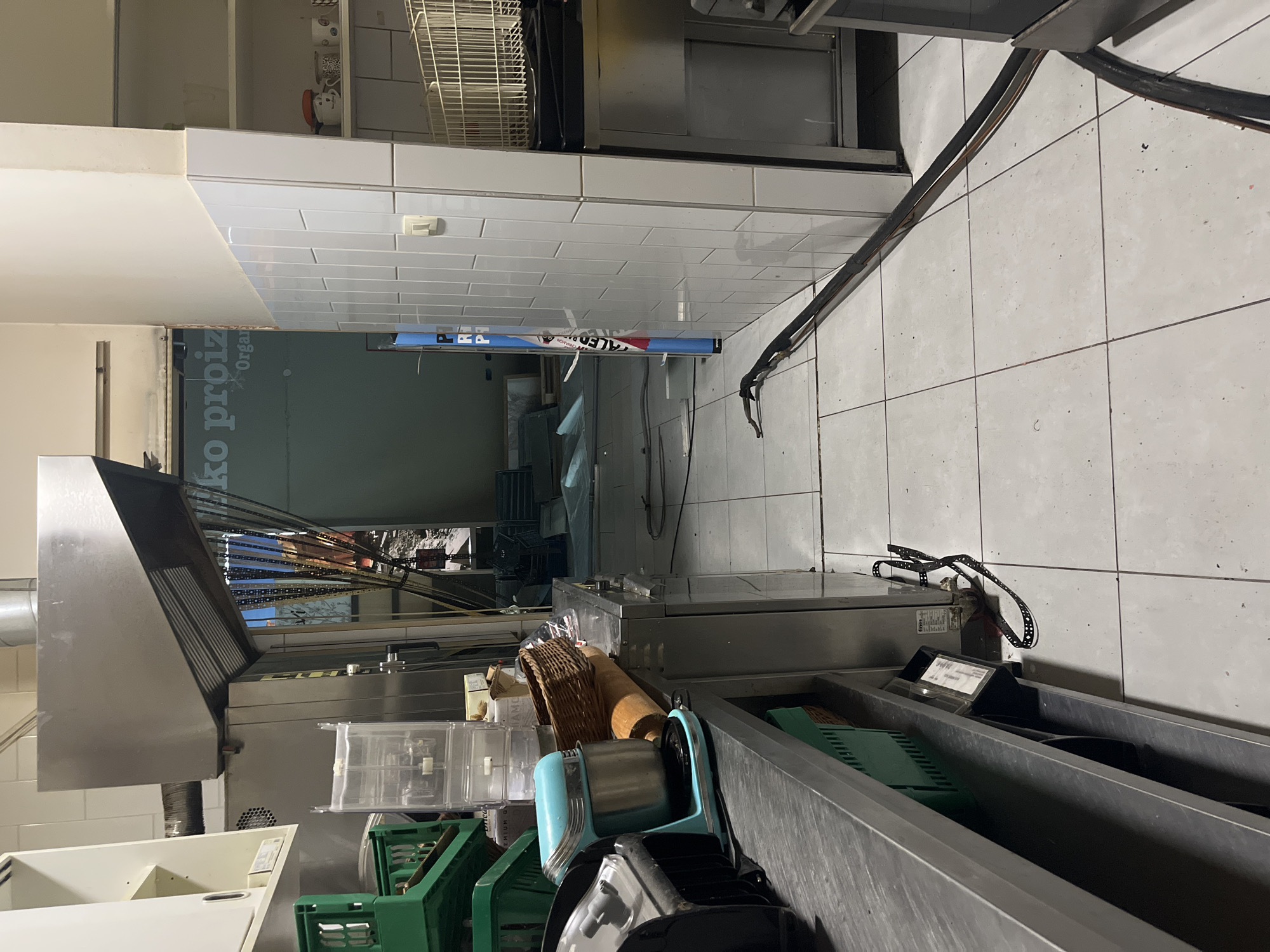 The image depicts the interior of what appears to be a commercial kitchen, likely in Budva, Montenegro, judging from text visible in a blurred background. The kitchen features tiled walls and flooring. Various pieces of commercial kitchen equipment are present, including stainless steel counters, a ventilation hood, and dish racks. Several green plastic crates and a blue food slicer are on the counter. A wicker basket and other kitchen supplies are scattered. The lighting is artificial and somewhat dim. The overall impression is of a functional, working kitchen environment. No people are visible in the image.