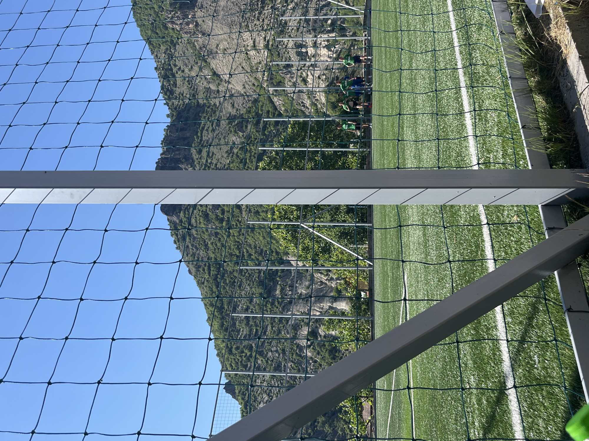 A view through a fence and a metal structure of a soccer field in Bukovo, Bulgaria. The field has artificial turf and white lines. A group of people wearing green shirts are visible along the field. In the background, a mountain rises behind the field against a clear blue sky. Grass and concrete border the field on the right side of the image.