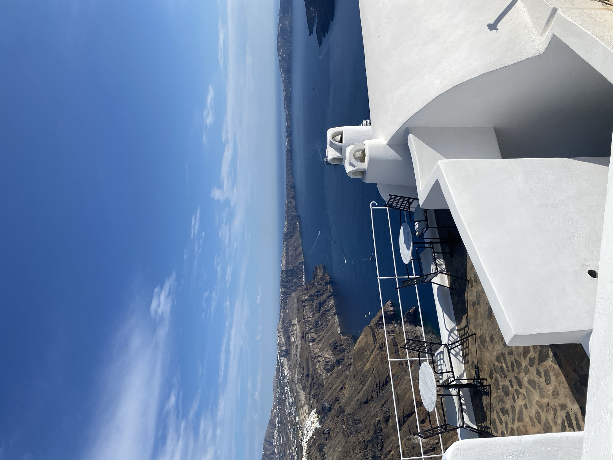 This media file depicts a high-angle view of a scenic coastline and a white, curved building, likely a cafe or restaurant, in Santorini, Greece. The scene is bathed in bright sunlight, suggesting a clear day, possibly mid-morning or early afternoon. The sky is a vibrant blue with scattered wisps of white clouds, indicating good weather.
The building, painted a stark white, features rounded architectural elements and several decorative white structures that resemble chimneys or ventilation shafts. In the foreground, outdoor seating is arranged on a patio paved with irregular, dark stones. Two small, round tables with black metal chairs are visible. A white railing separates the patio from the edge of the cliff.
Beyond the building and patio lies a dramatic caldera landscape. The deep blue sea stretches out to the horizon, dotted with a few small boats. The rugged, steep cliffs of the caldera rise from the water, showcasing layers of dark rock and sparse vegetation. Along the clifftop, a ribbon of white buildings, characteristic of Santorini's villages, is visible nestled against the dark terrain. The juxtaposition of the pristine white architecture, the deep blue sea, and the dramatic, rocky landscape creates a striking visual contrast.
There is no visible text in the image. The overall mood conveyed is one of serene beauty and luxurious relaxation, characteristic of the popular tourist destination.