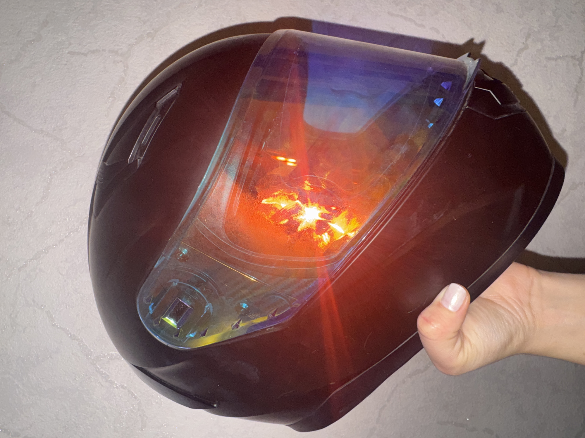 A hand with painted fingernails holds a dark red motorcycle helmet. The helmet has a tinted visor that reflects a bright orange and yellow light. The background is a textured, light-colored wall. The image is taken from a medium close-up angle. The location is not specified in the image.