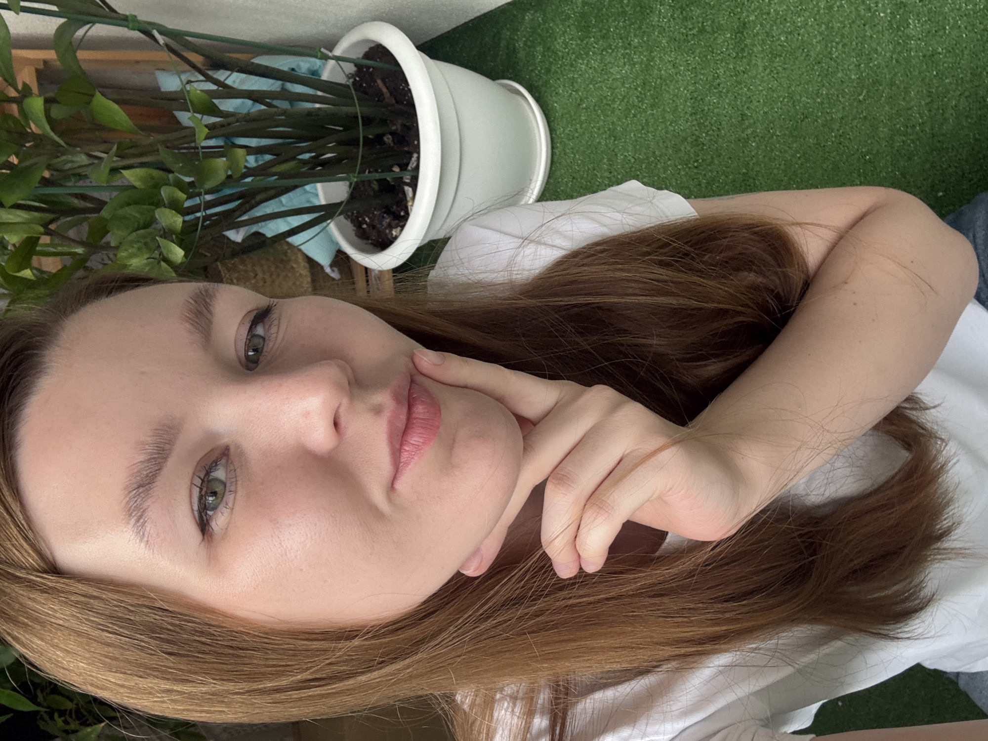 The image is a close-up selfie of a young woman with auburn hair and light skin. She is wearing a white t-shirt and has her right hand on her chin, with her fingers delicately touching her lips. Her eyes are green, and she appears to be looking directly at the camera with a slight smirk. In the background, a potted plant with dark green leaves is visible, along with a white plastic pot and a blue cloth. The ground appears to be covered in artificial turf. The image does not contain any specific elements that would indicate its location within the city of Izhevsk, Russia.