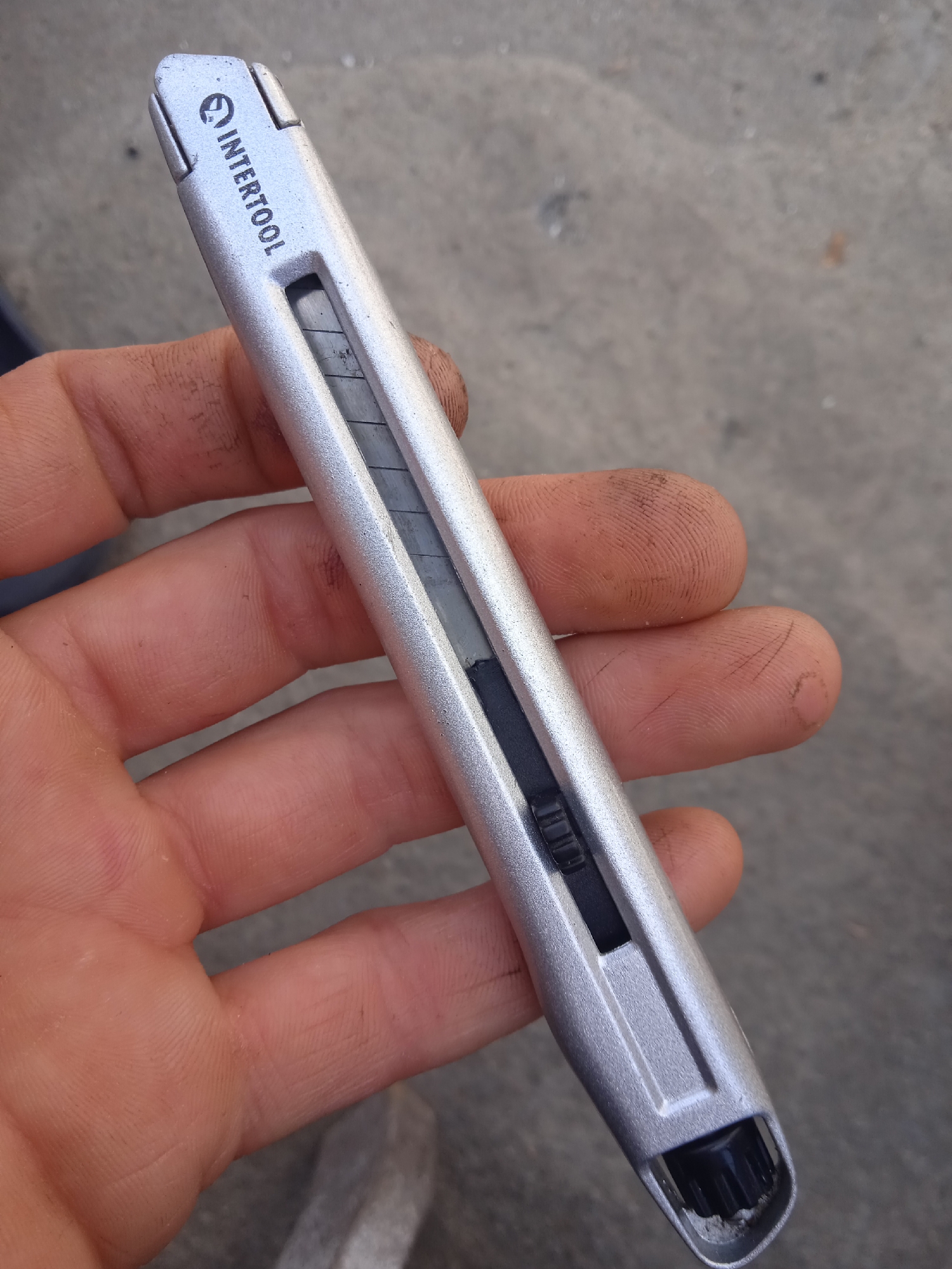 A hand with dirty fingers holds a silver utility knife. The knife has a black slider mechanism to extend and retract the blade, which is partially visible and appears to be segmented. The brand name "INTERTOOL" is visible in black lettering at the top of the knife. The background is a rough, greyish surface, possibly concrete or pavement.