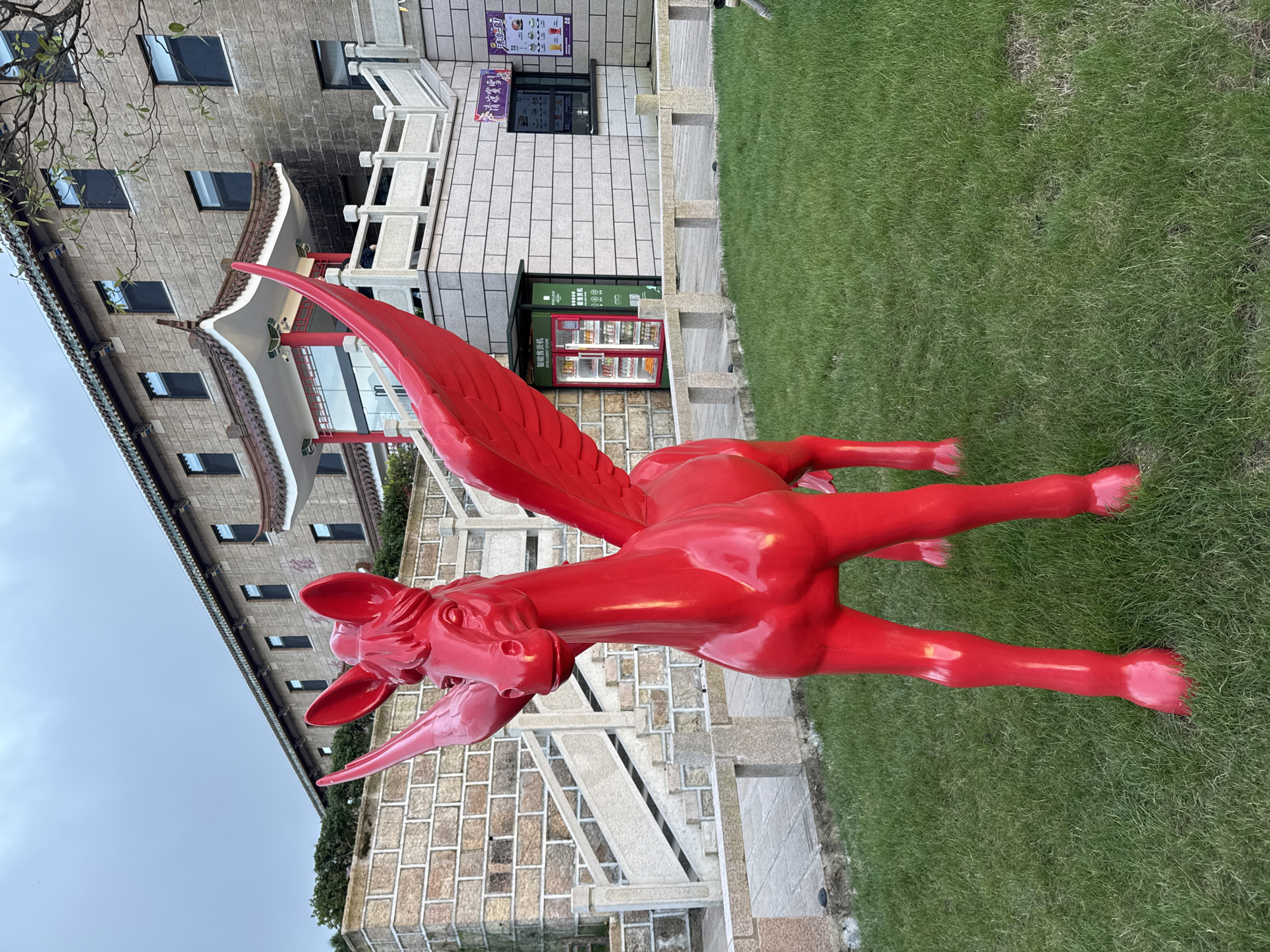 A bright red, glossy, winged horse statue stands on a well-maintained green lawn in front of a stone building with a traditional Chinese architectural style. The building has a multi-tiered, curved roof with green tiles and a facade of rough-hewn stone blocks. White stone railings and stairs lead up to the building. To the right of the building, there is a modern, green vending machine. The sky is overcast. The statue is the most prominent object in the foreground.