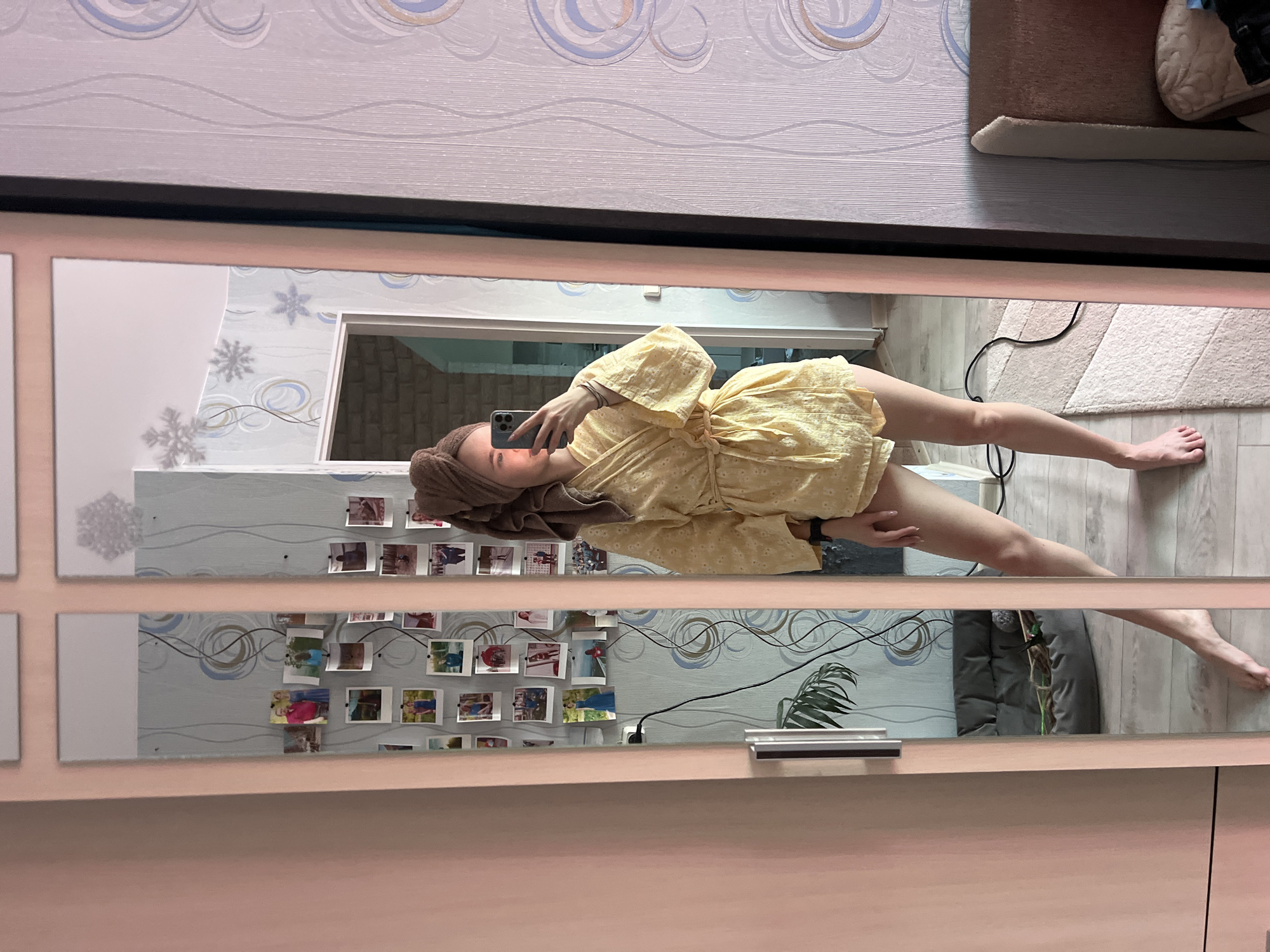 A woman in a yellow dress and brown towel on her head takes a selfie in a mirror. The mirror hangs on a pink wall in what appears to be an apartment. The wall behind her has a patterned wallpaper with swirling designs and affixed photographs. A potted plant sits on a table next to the mirror. The setting is a room in Chaikovsky, Russia.