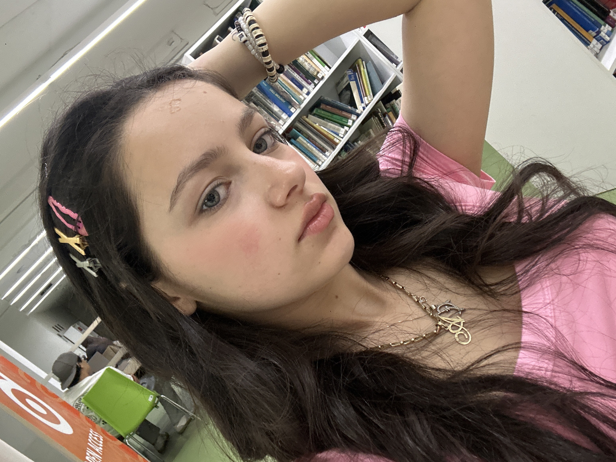 The photograph presents a young woman with long, dark brown hair and fair skin. She has blue eyes, and her lips appear to be lightly tinted with gloss. Hair clips are visible on the left side of her head. She is wearing a pink top and a gold necklace with an ornate pendant. Her arm is raised, with a beaded bracelet visible on her wrist. In the background, there are bookshelves filled with books, suggesting a library setting. Other people are visible in the background, as well as green seating and an orange sign. There is no clear indication of Yerevan, Armenia in the image.
