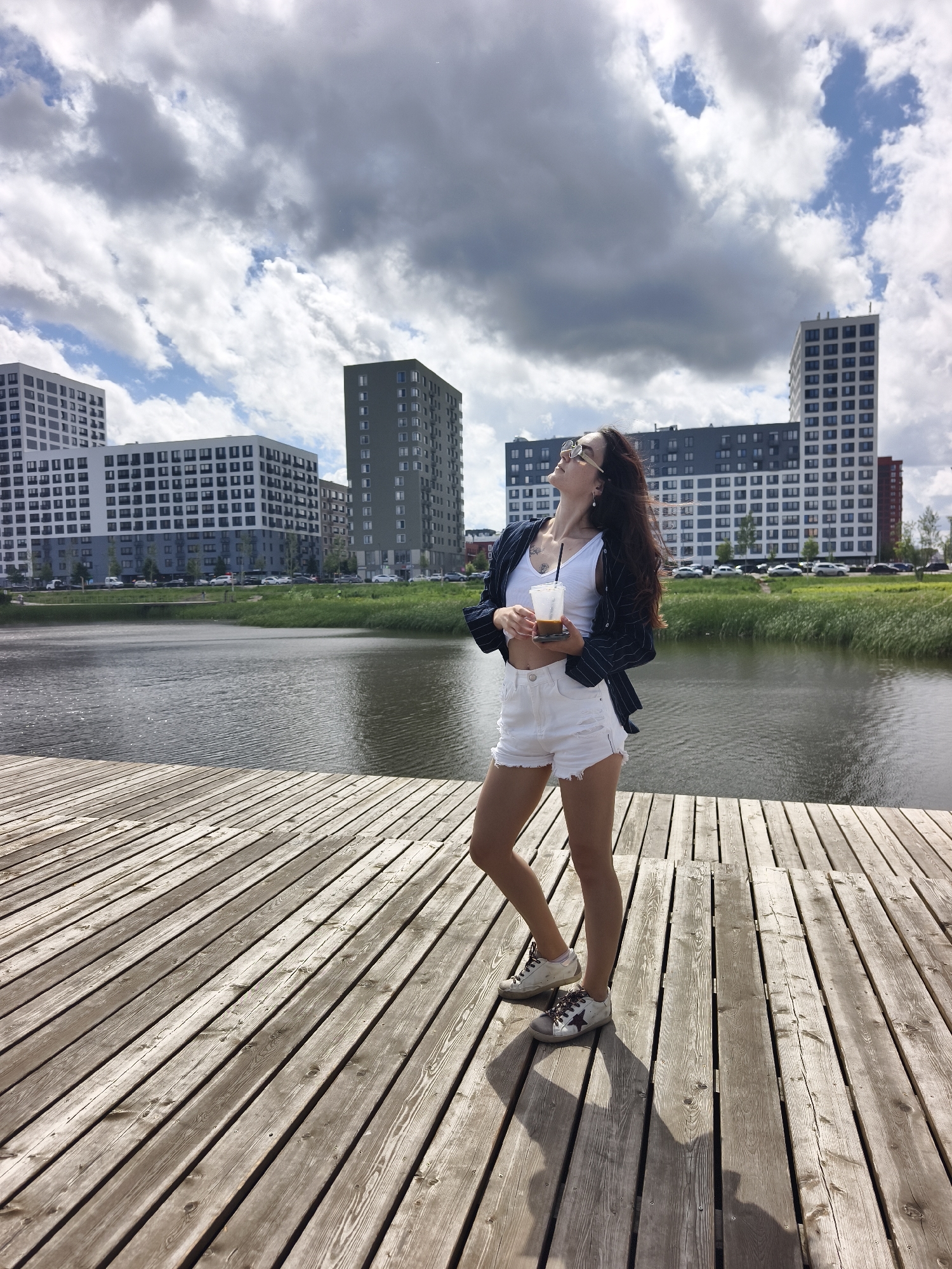 In Tyumen, Russia, a young woman stands on a wooden pier, holding a to-go cup of coffee. She is dressed in white shorts, a white tank top, and a striped blazer. She wears sunglasses and her hair is long and dark, blowing in the wind. Behind her, a body of water is visible, with modern apartment buildings in the distance. The sky is filled with dramatic clouds.