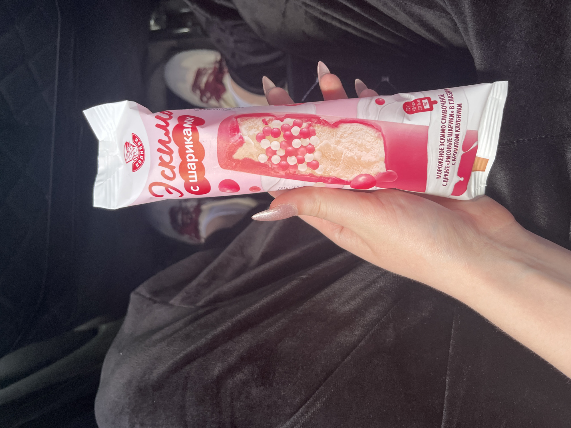 A person is holding a pink and white popsicle wrapper with an ice cream image on it. The wrapper has Russian text. The person's hand, with long, light-colored acrylic nails, is holding the wrapper. The person is wearing dark, loose-fitting pants. The background shows a dark interior, possibly a car, with blurred feet wearing white shoes with red accents visible. The location is identified as Novaya Otradovka, Russia.
