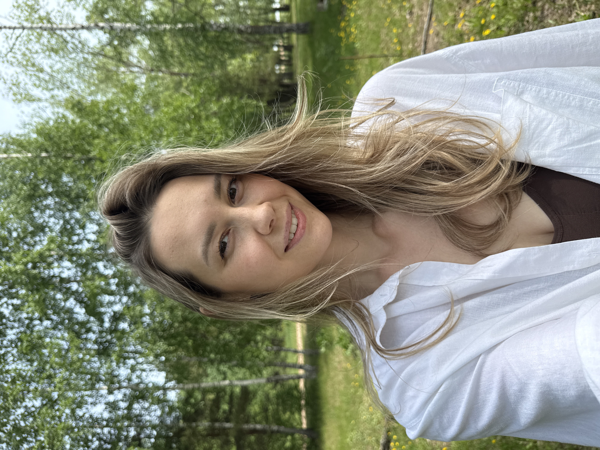 A woman poses outdoors in Noviy, Russia. She is wearing a white shirt over a brown tank top. Her hair is light brown and wavy. The background features trees and grass.