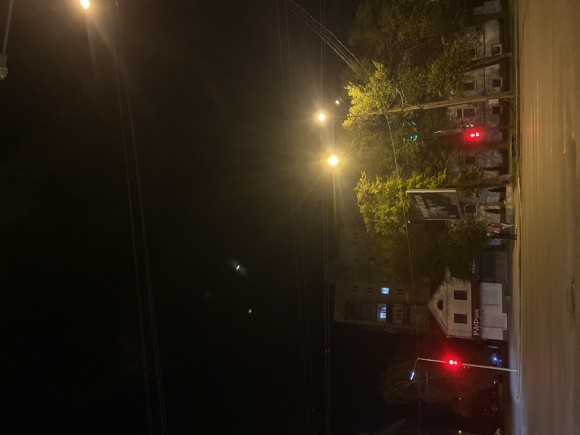 A nighttime scene in Poltava, Ukraine. The view is along a street at night. The street lights illuminate trees and nearby buildings. A traffic light is visible in red, and a pedestrian stands on the sidewalk near a storefront with Cyrillic script. Overhead wires are also visible.