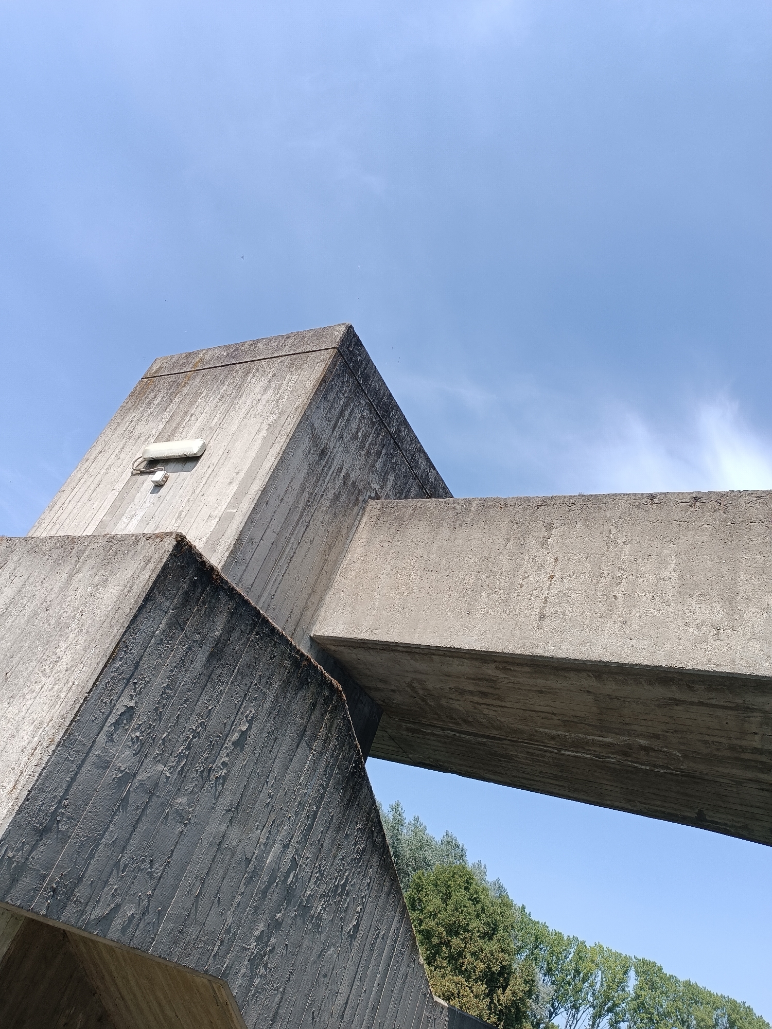 The image shows a low-angle, outdoor shot of a Brutalist concrete structure against a clear blue sky with faint wisps of clouds. The concrete elements are angular and geometric, with visible texture from the formwork used during construction. One element is a large, angular beam intersecting with another prominent concrete surface. On the upper portion of one of the concrete faces, a white light fixture with a smaller box-like attachment is visible. In the background, a line of lush green trees forms a natural border against the sky. The scene appears to be a public space, possibly a park or a plaza, in the city of Casalmaggiore, Italy, given the context.