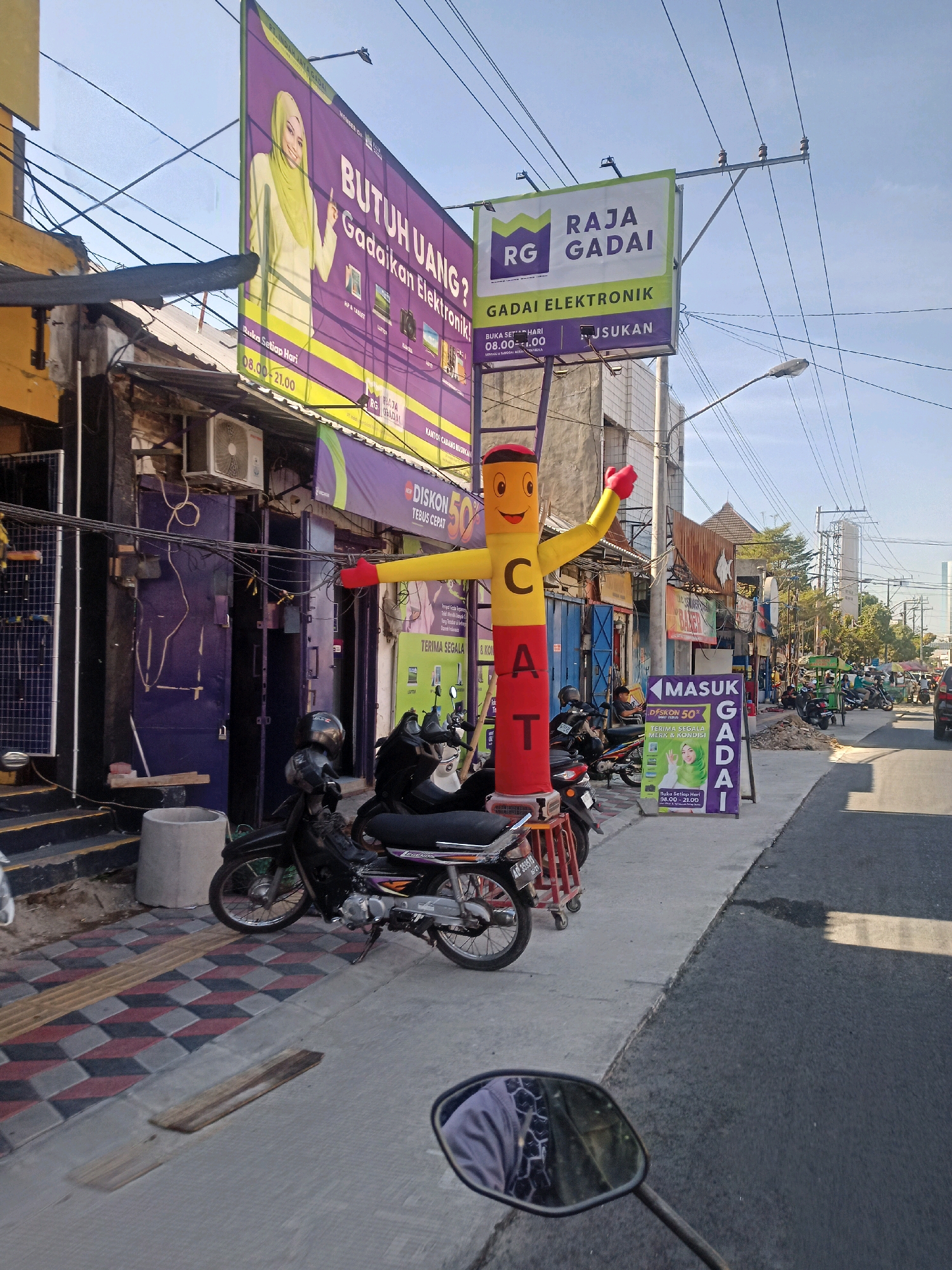 The image depicts a street scene in Surakarta, Indonesia, likely a commercial area. Prominently featured is a large purple and yellow billboard advertising "BUTUH UANG? Gadaikan Elektronik!" (Need Money? Pawn Electronics!), showing a woman in a hijab. Adjacent to it is a green and white sign for "RAJA GADAI ELEKTRONIK" (King Pawn Electronics). A large inflatable tube man, red and yellow with a smiley face, is positioned on the sidewalk.
The sidewalk is paved with patterned tiles and concrete. Several motorcycles are parked along the sidewalk. A building with dark purple doors and air conditioning units is visible behind the inflatable man. Further down the street, other shops and businesses are present, along with more parked motorcycles and some moving vehicles. Utility wires are strung across the scene, and a street light is visible. The sky is clear and blue, suggesting a sunny day. The overall impression is that of a busy, everyday street in an Indonesian city.